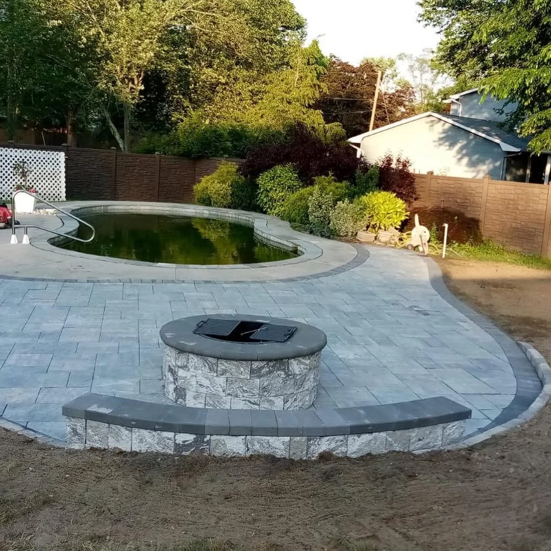 Backyard with empty swimming pool, surrounding patio, raised fire pit with seating, lush bushes, and a neighbor's house in the background.