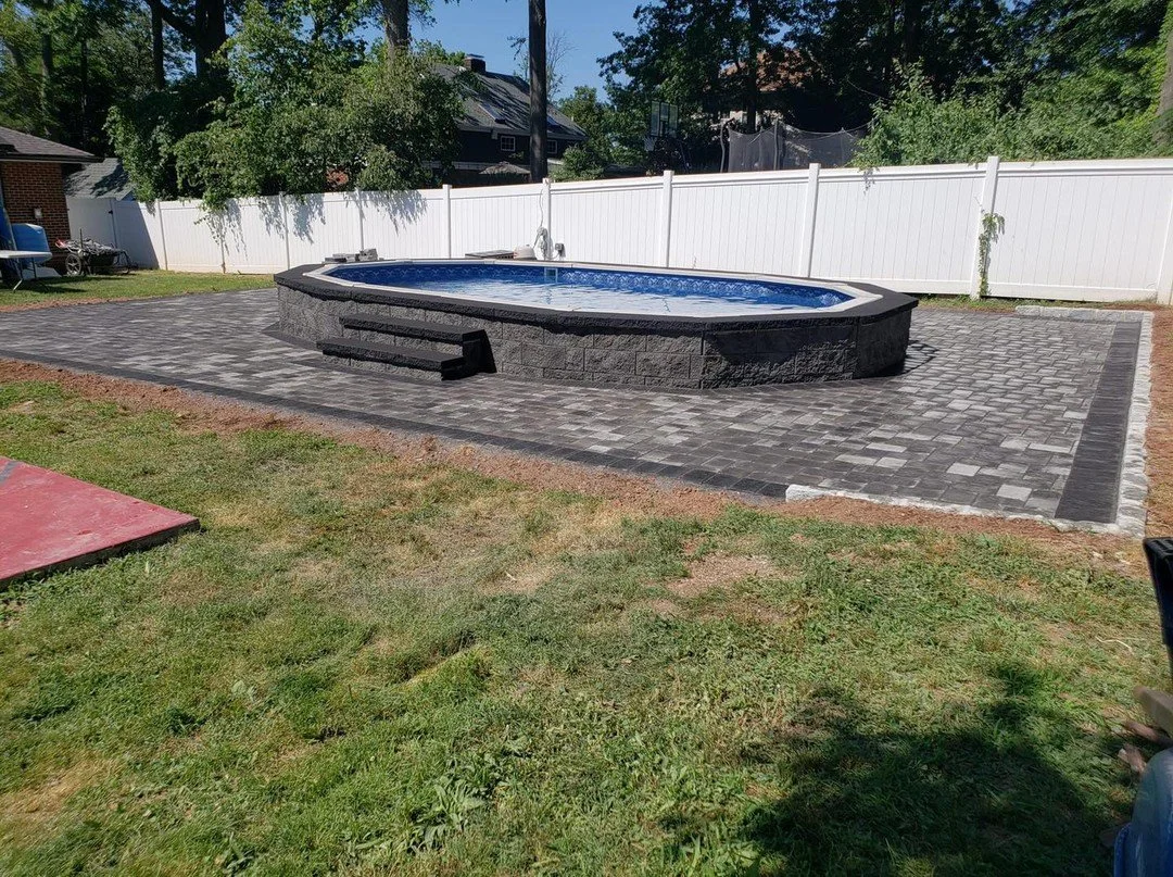 Above ground backyard swimming pool with a dark stone exterior, surrounded by a brick paver patio, in a fenced backyard with trees and neighboring houses.