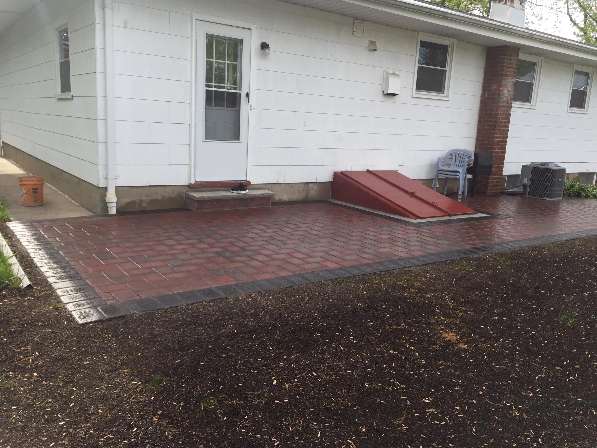 Back patio with brick pavers, white house exterior, door with shoe on porch step, gray plastic chairs, a grill, and an air conditioning unit.