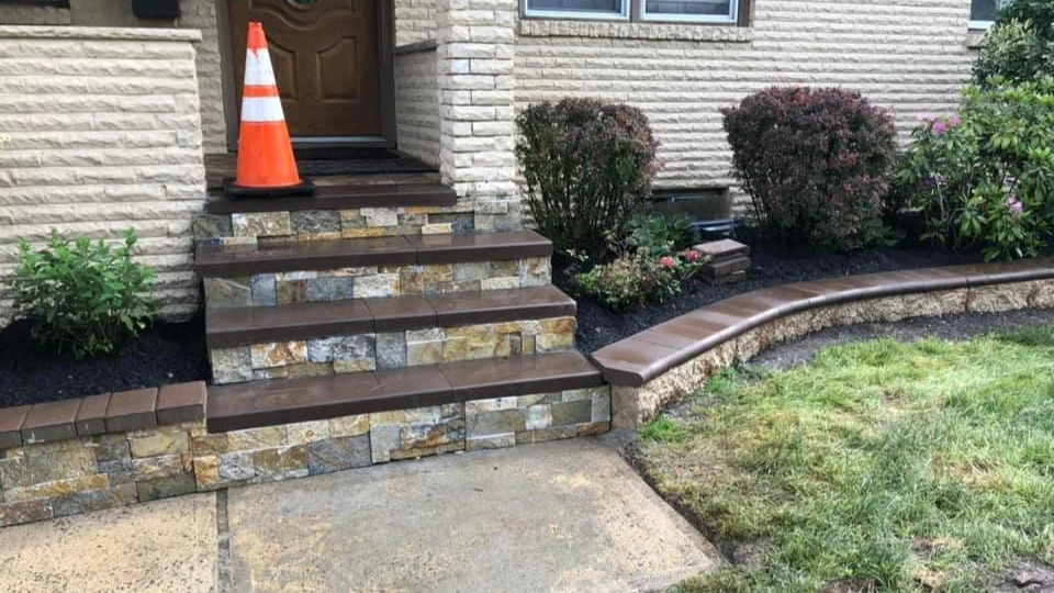 Stone steps with brown treads leading up to a house entrance, with an orange traffic cone on top of the steps. There are landscaped bushes and a curved stone retaining wall with a brown cap, surrounding a grassy area.