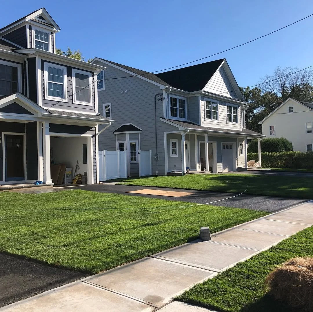 Newly constructed gray multi-story house with white trim, large windows, gabled roof, and a front porch with columns, surrounded by a green lawn and a paved walkway, under a clear blue sky.