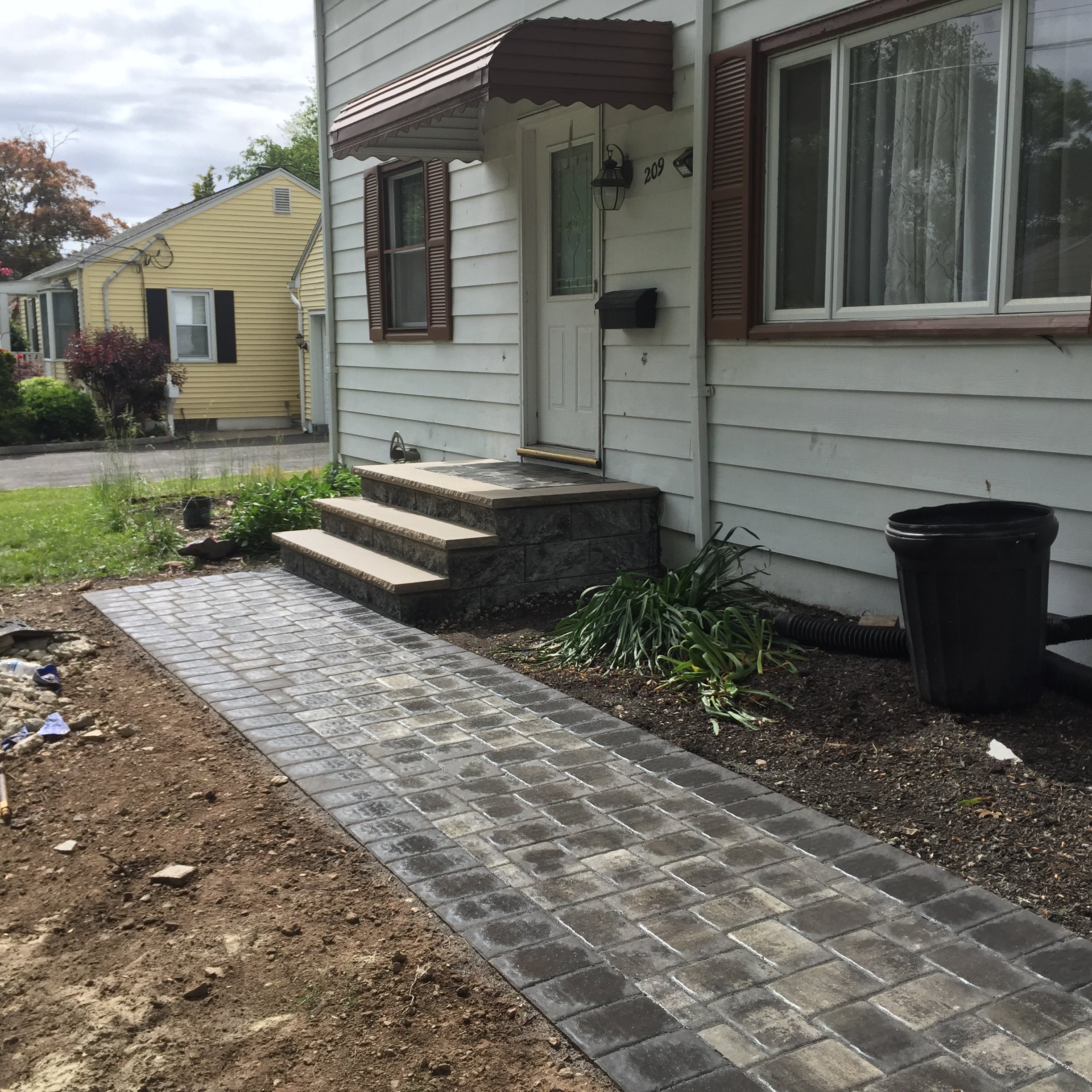 Front porch with stone stairs leading to a white house with brown shutters and a small garden bed with plants, a newly laid brick walkway, and overcast sky.