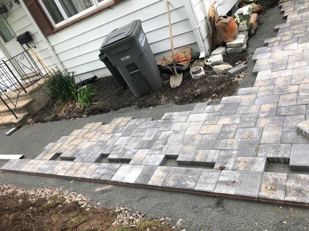 Newly paved section of a walkway with gray interlocking bricks. Construction materials and tools, including a shovel and bags of material, are visible along the gravel border to the right. A house with white siding and a small garden with green plant