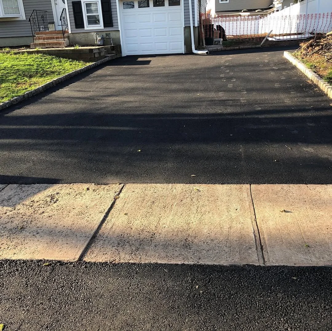 Newly paved driveway in front of a house with a white garage door, adjacent to a concrete sidewalk and a small green lawn.