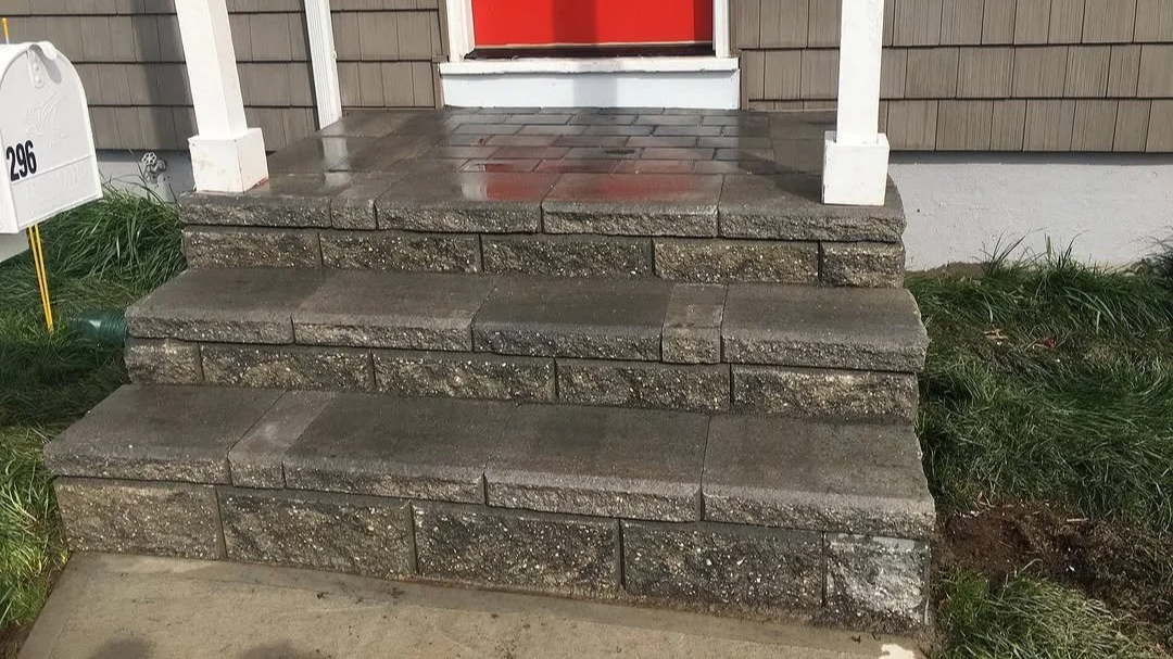 Concrete steps leading to a front porch with a red door and white supports, surrounded by grass and a gray siding house.