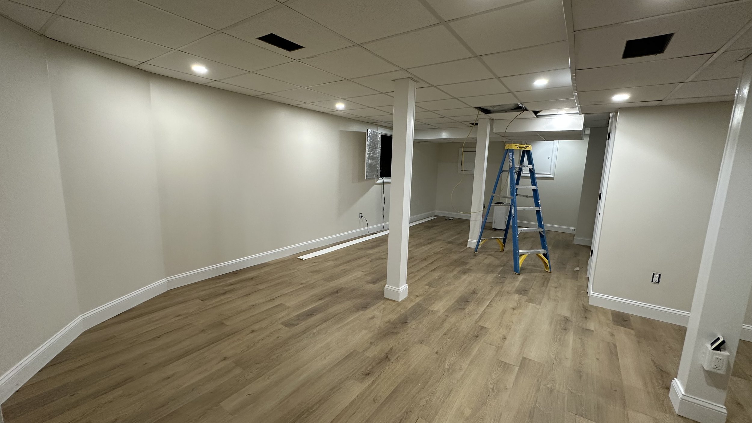 Undeveloped basement with beige walls, light wood flooring, drywall ceiling with recessed lighting, a blue and yellow ladder, and an open electrical panel.