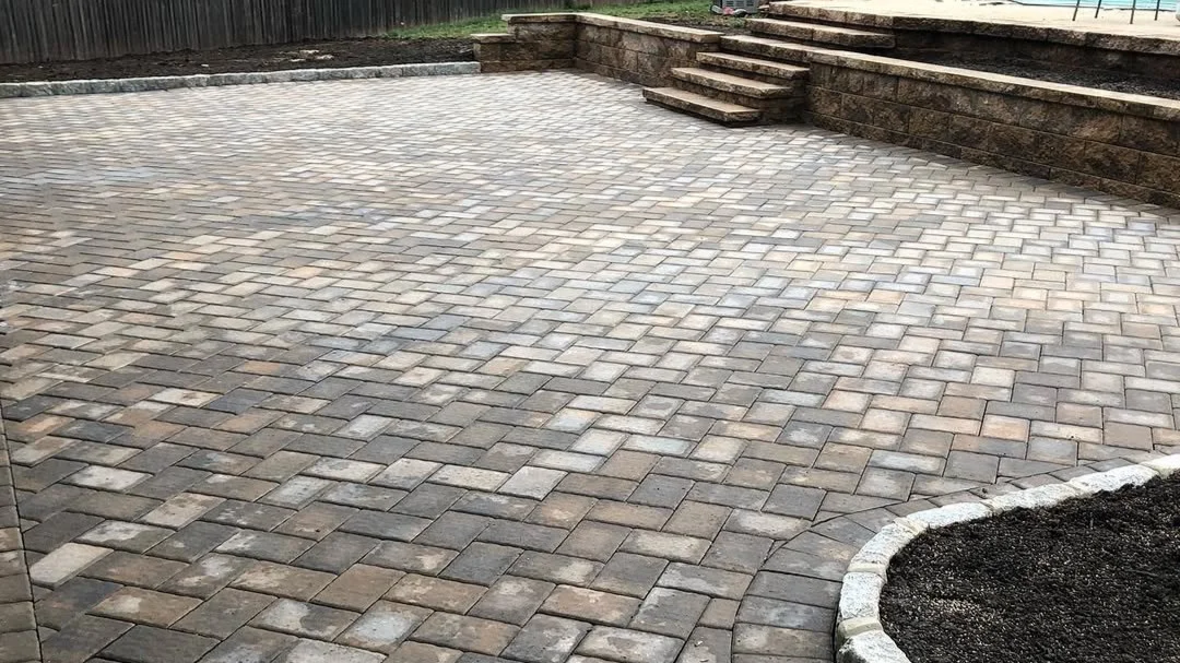 Photo of a paved backyard area with brick pavers, a stone retaining wall, and a small set of stairs leading up to another level.