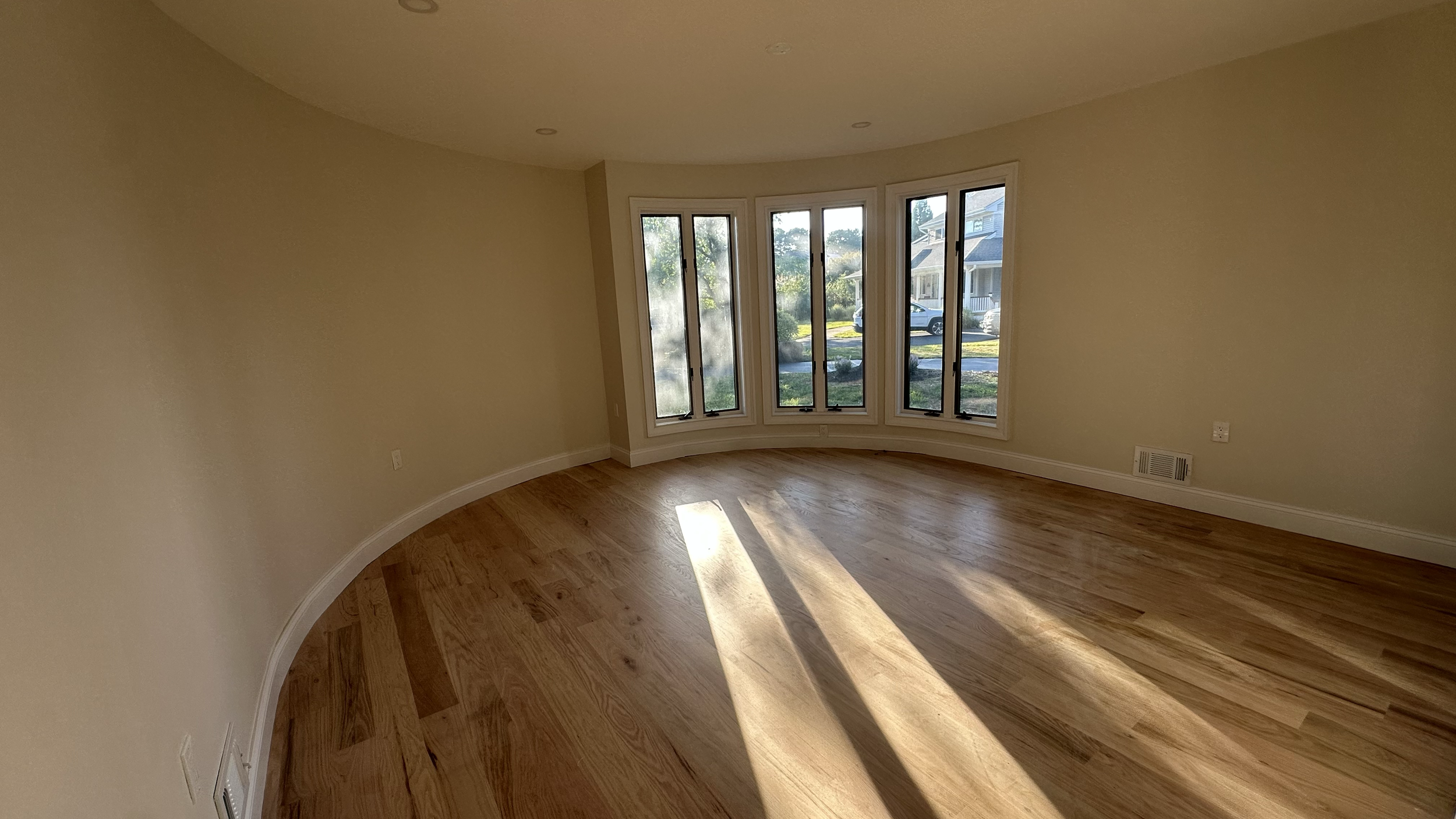 Empty room with hardwood floor, cream-colored walls, and large bay window letting in sunlight, with a view of neighboring houses outside.