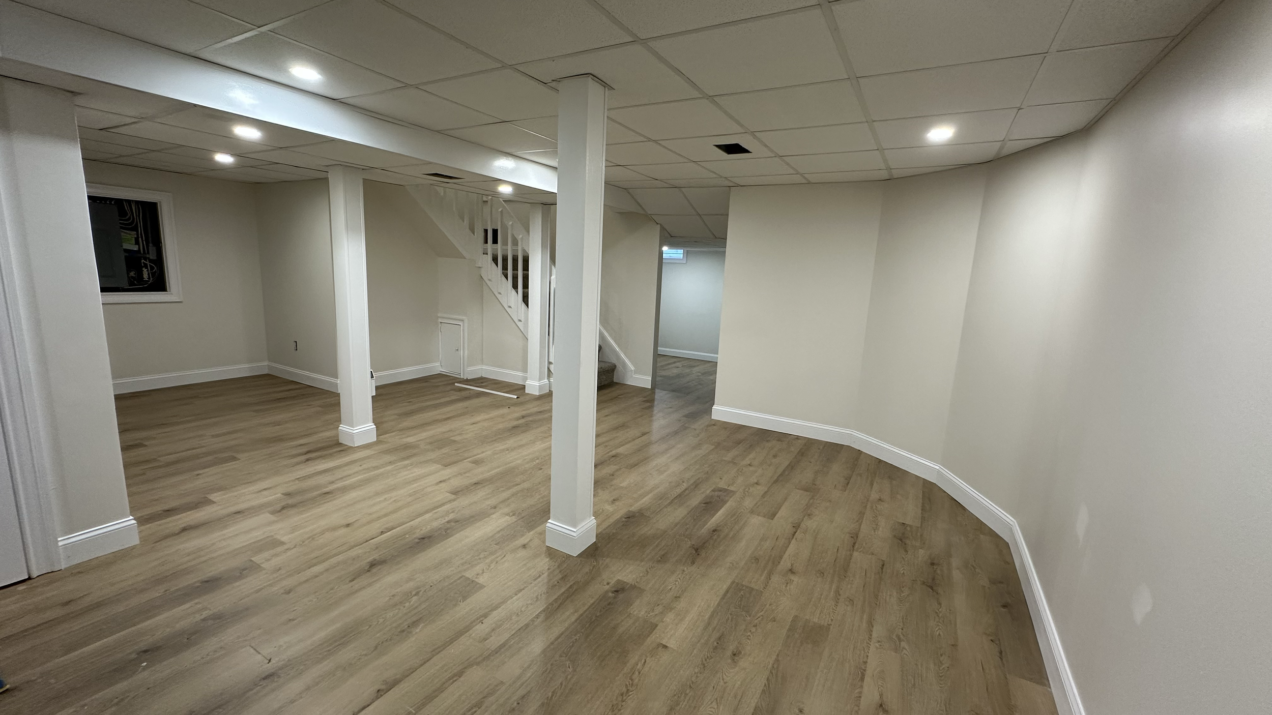 Empty basement room with wooden flooring, white walls, ceiling lights, and a staircase with white railing.