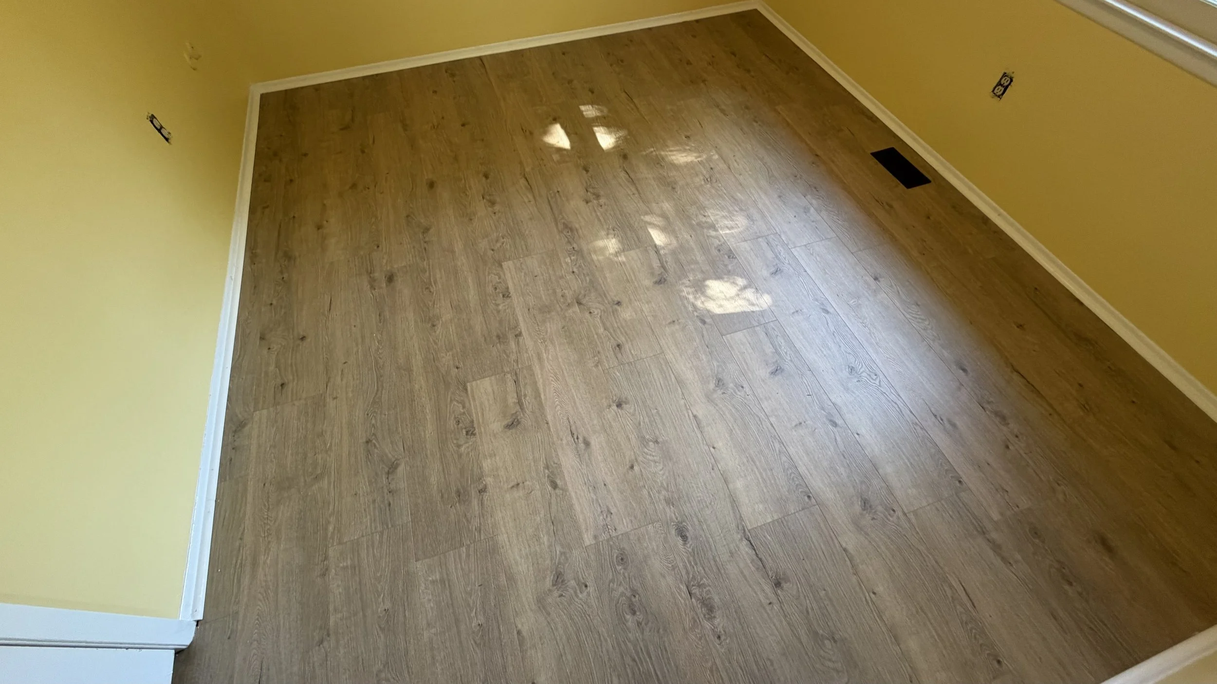 Empty corner of a room with yellow walls, wooden flooring, white baseboards, and a power outlet.