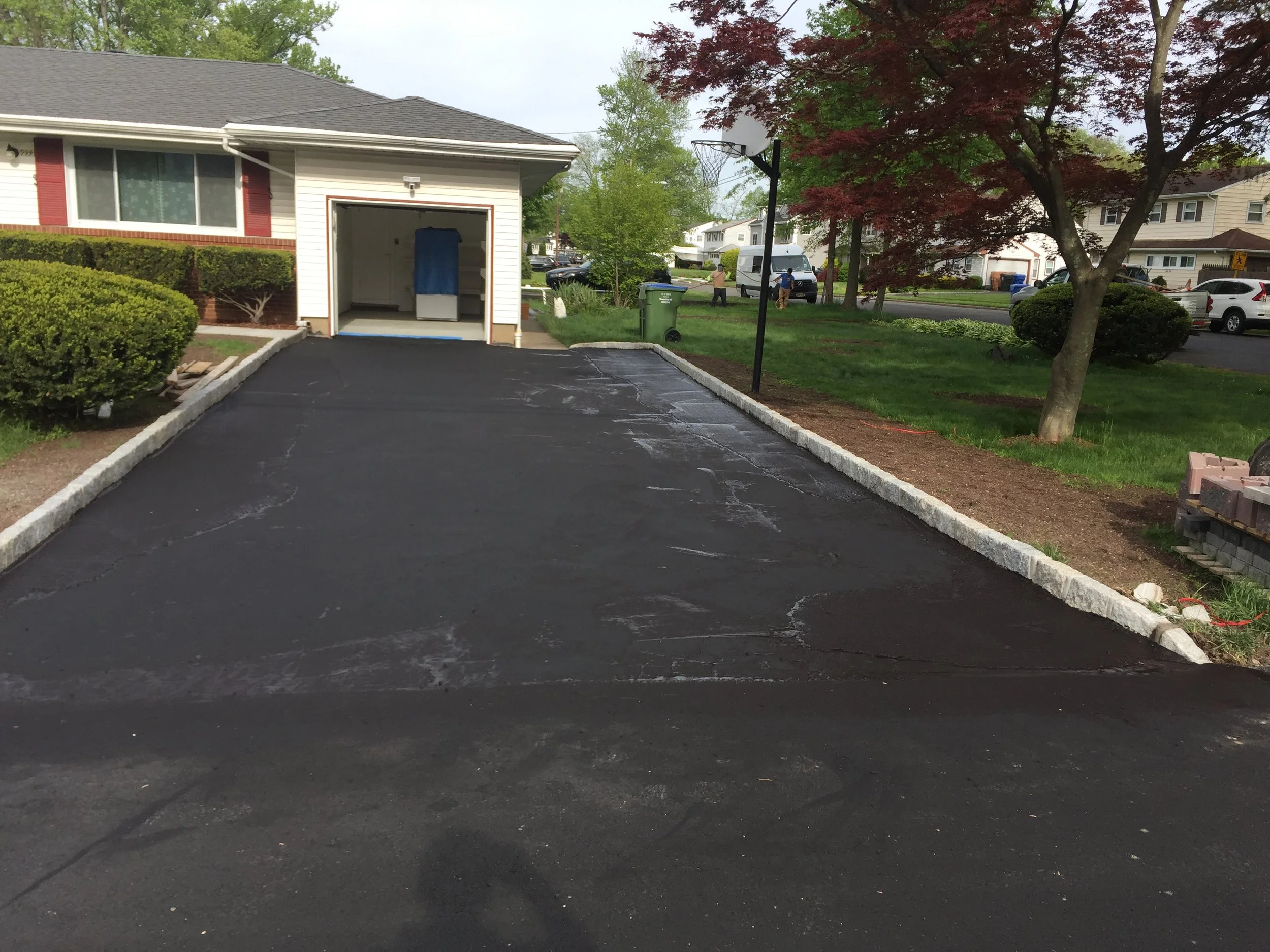 A freshly paved black asphalt driveway with a concrete border, leading to a garage with a blue cover inside. There are green bushes and a small red-leafed tree on the right side, with a basketball hoop mounted on a pole near the tree. In the backgrou
