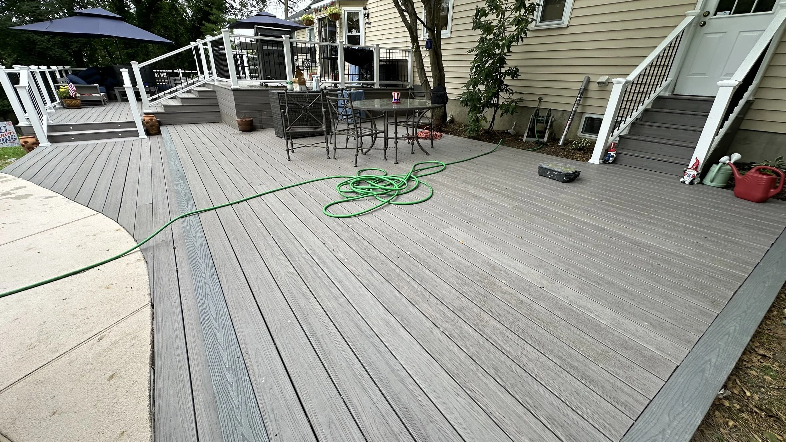 A wooden deck outside a house with patio furniture, a green garden hose, and garden tools; some umbrellas are open, and decorative items are visible.