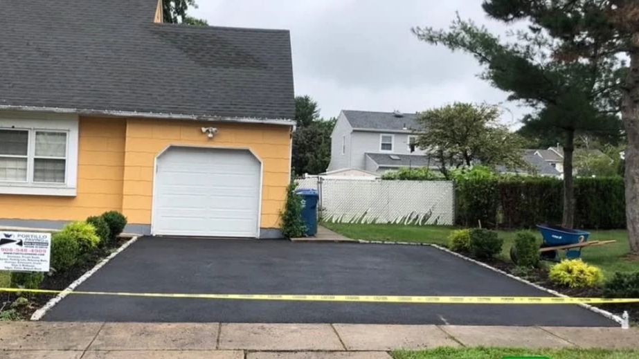Recently paved driveway in front of yellow house with white garage door, surrounded by small bushes and yellow tape, with a hand truck and garden tools on the lawn to the right.