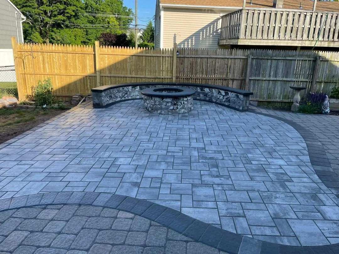 Fully paved backyard with decorative stone fire pit and curved seating wall, fenced backyard with neighboring house and trees in the background.