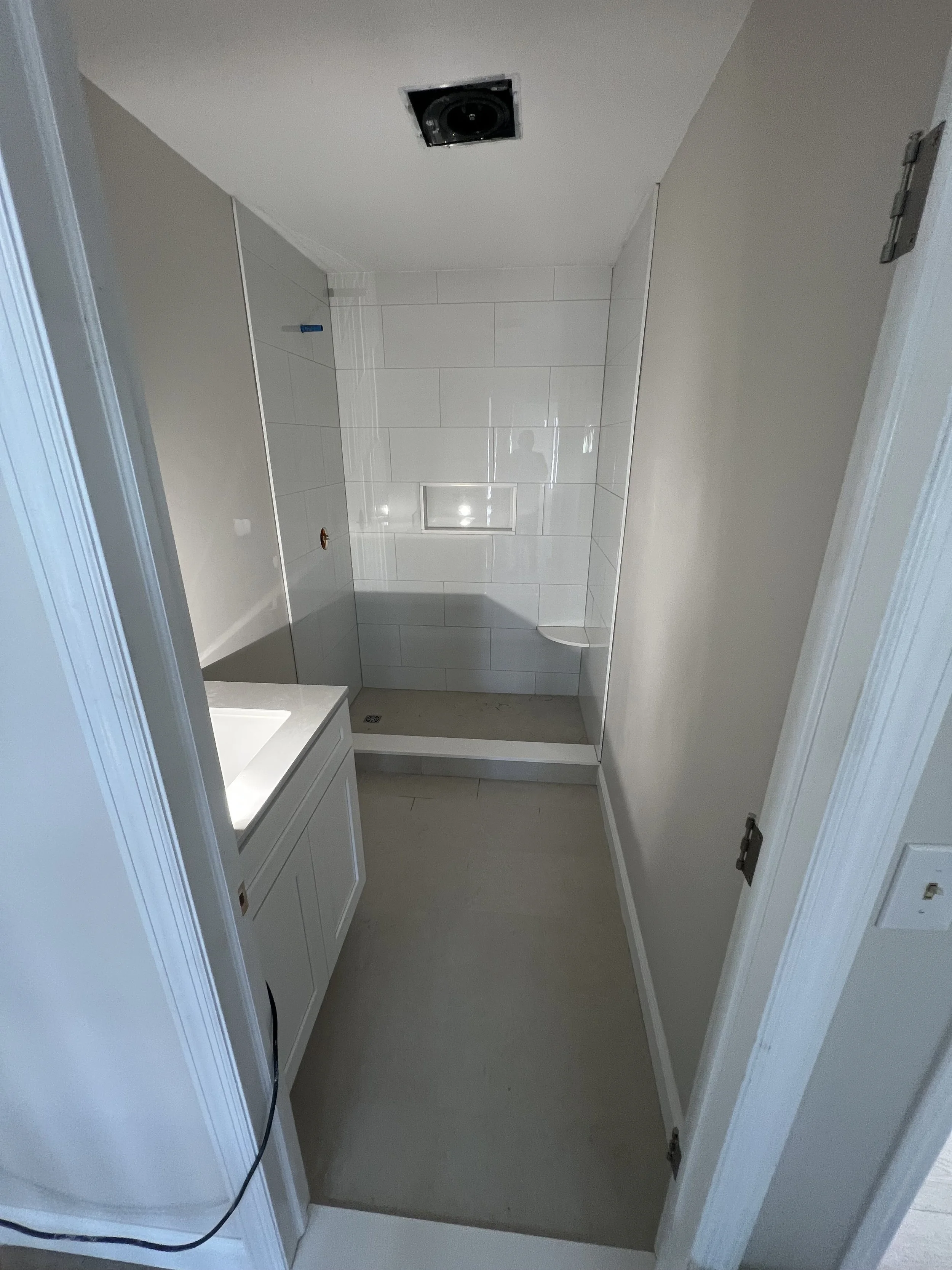 A bathroom with white tiles on the shower wall, a built-in niche, a bathtub, and a small vanity with a sink and cabinet. The ceiling has an opening for a light fixture or vent. The floor is a light beige color, and there is a visible black power cord hanging from the vanity.