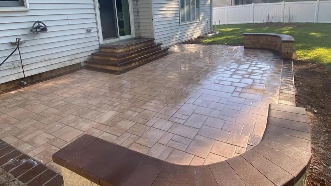 Newly constructed brick patio with steps leading to house and a curved brick bench along the edge, surrounded by a lawn and a white fence.