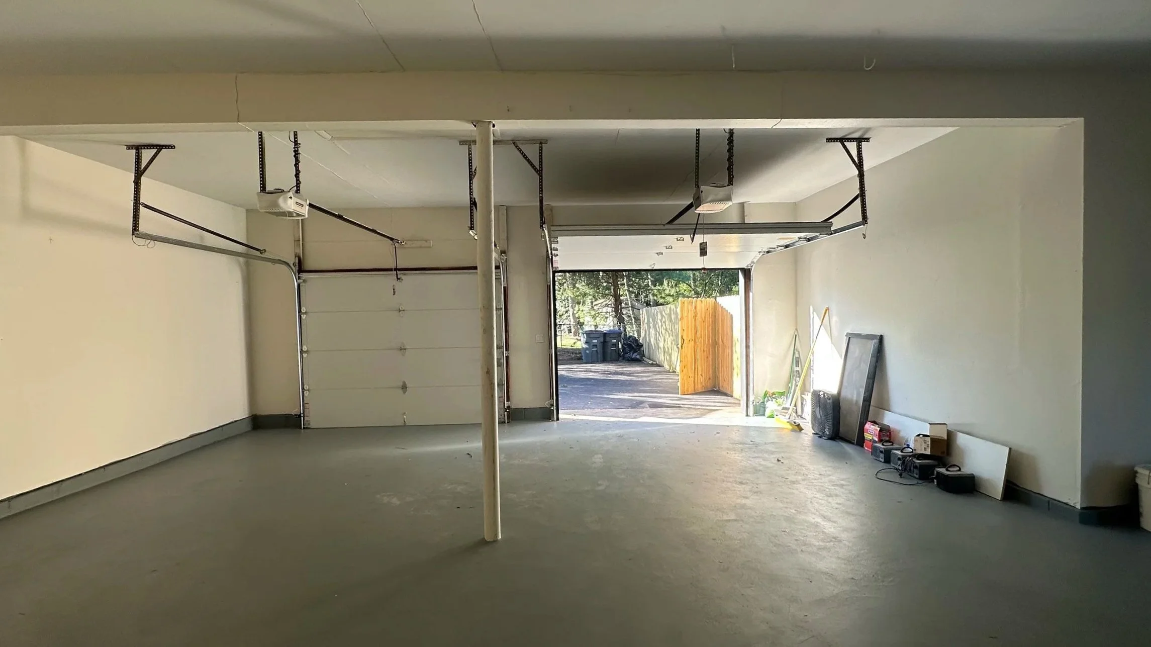 Empty garage with raised overhead door, concrete floor, and various tools and items along the right wall, open to outside with trees and trash bins visible.