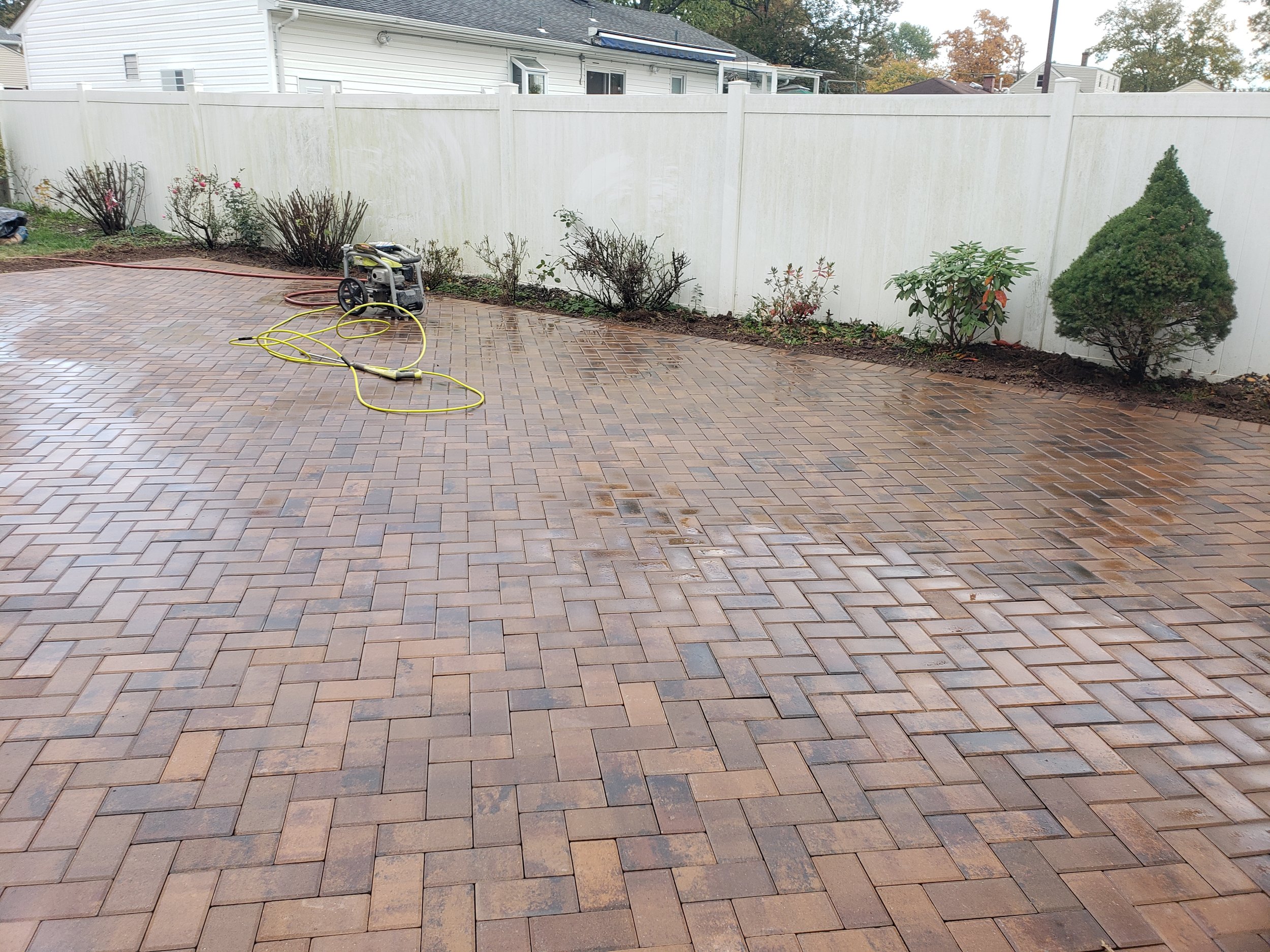Wet brick patio with a yellow hose and pressure washer, bordered by a white fence and garden plants.