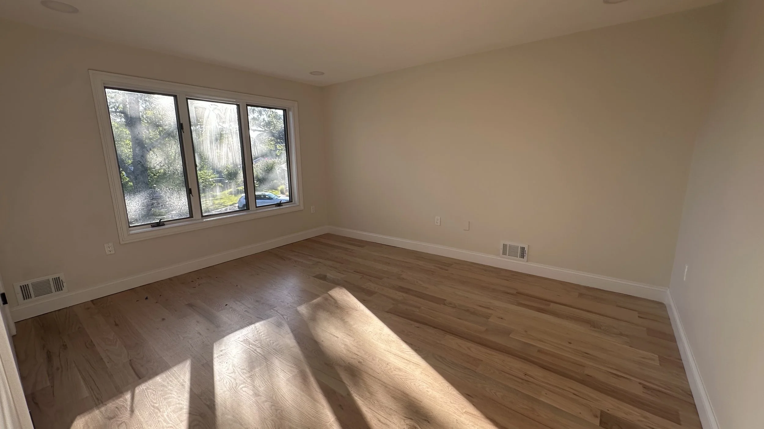 Empty room with hardwood floors, a large window with a view of trees and a car, and white walls.