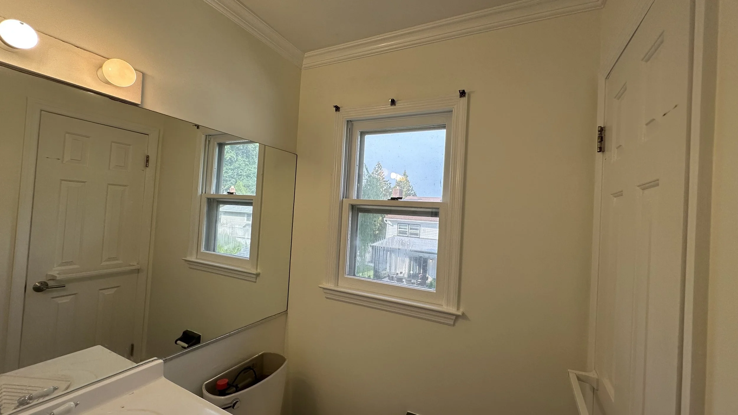 A small bathroom with cream-colored walls, a large mirror, a double-hung window, and a closed white door. An overhead light fixture with two bulbs is visible. Parts of a washing machine and small collection of items in an open laundry basket are seen at the bottom of the image.