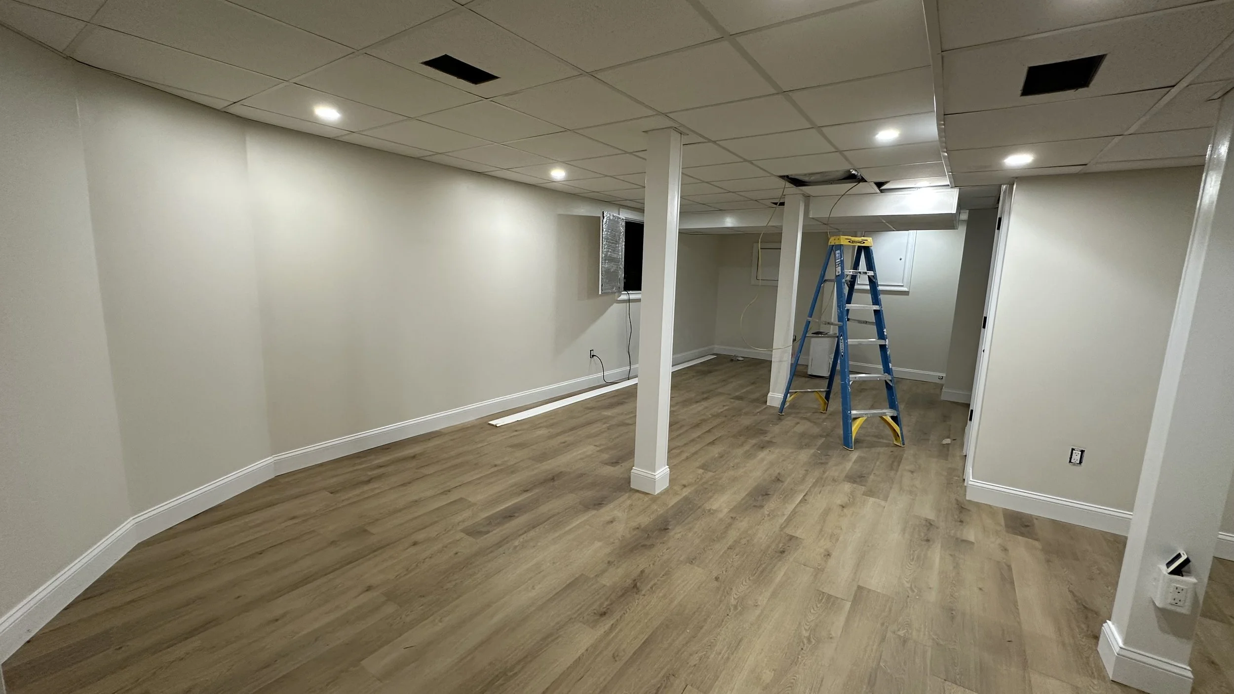 Undeveloped basement with wood flooring, white walls, ceiling with recessed lighting, a small window with duct tape, a blue ladder, and some construction tools.