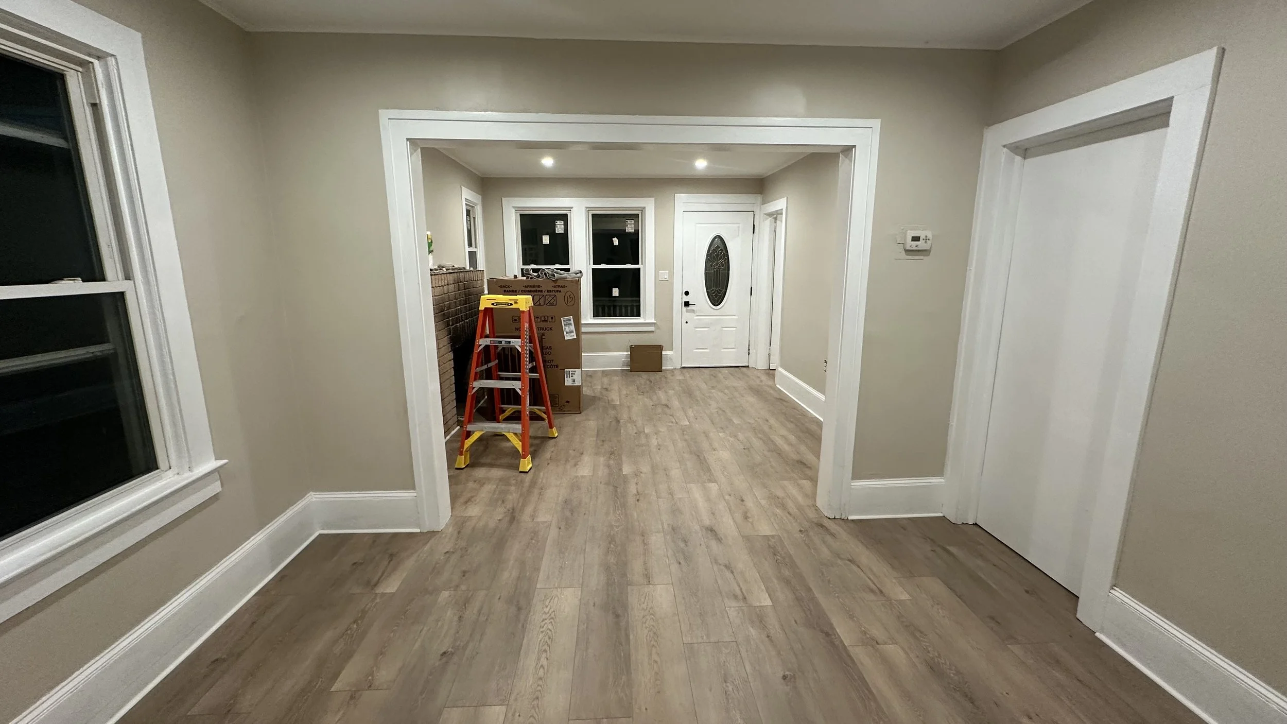 Empty living room with beige walls, white trim, and hardwood floors, leading to a foyer with a door and windows, containing a ladder and a large cardboard box.
