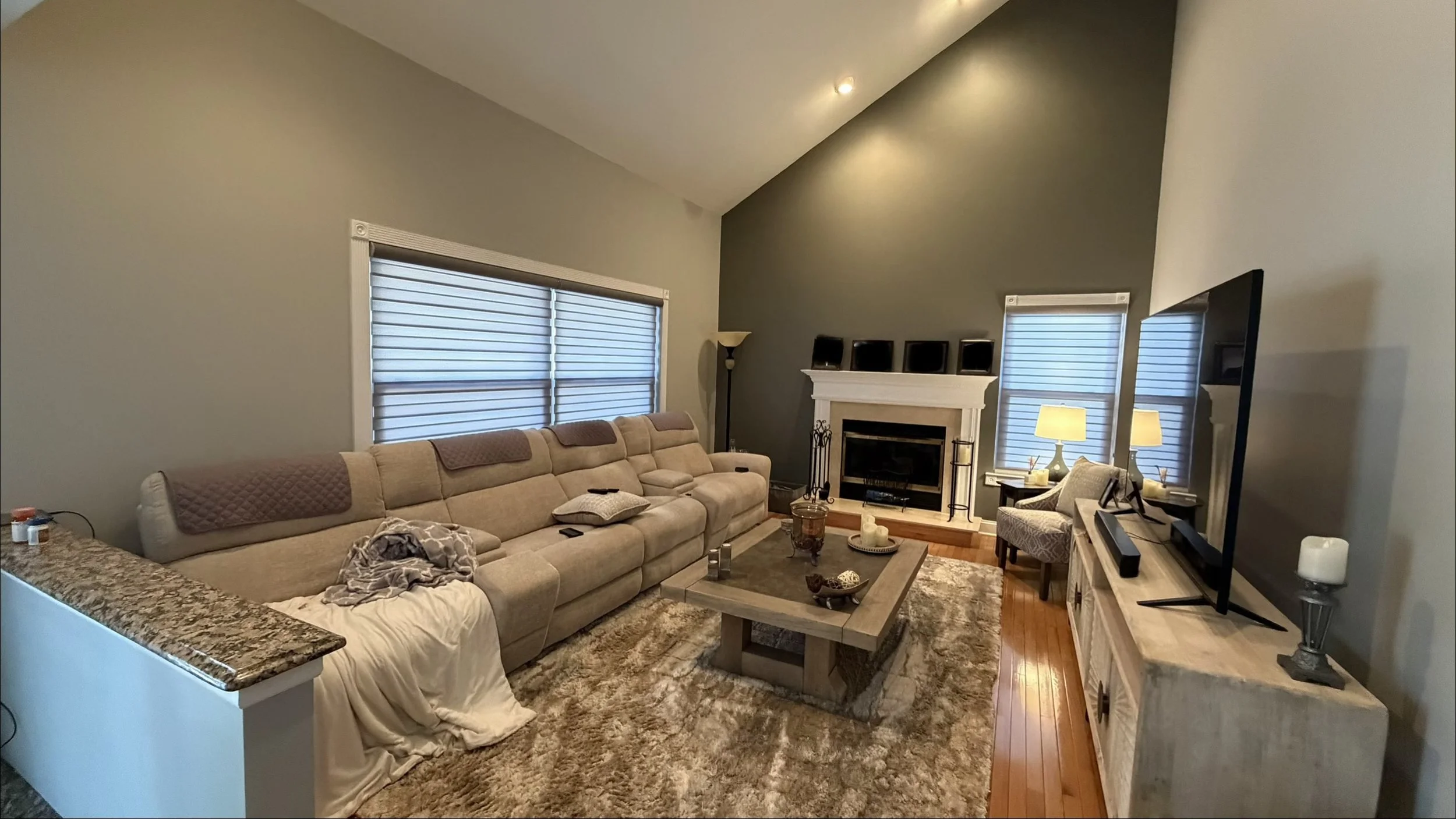 Living room with beige sectional sofa, fireplace with mantle, TV on a stand, windows with blinds, armchair, coffee table with decor, lamps, area rug, and decorative candles.