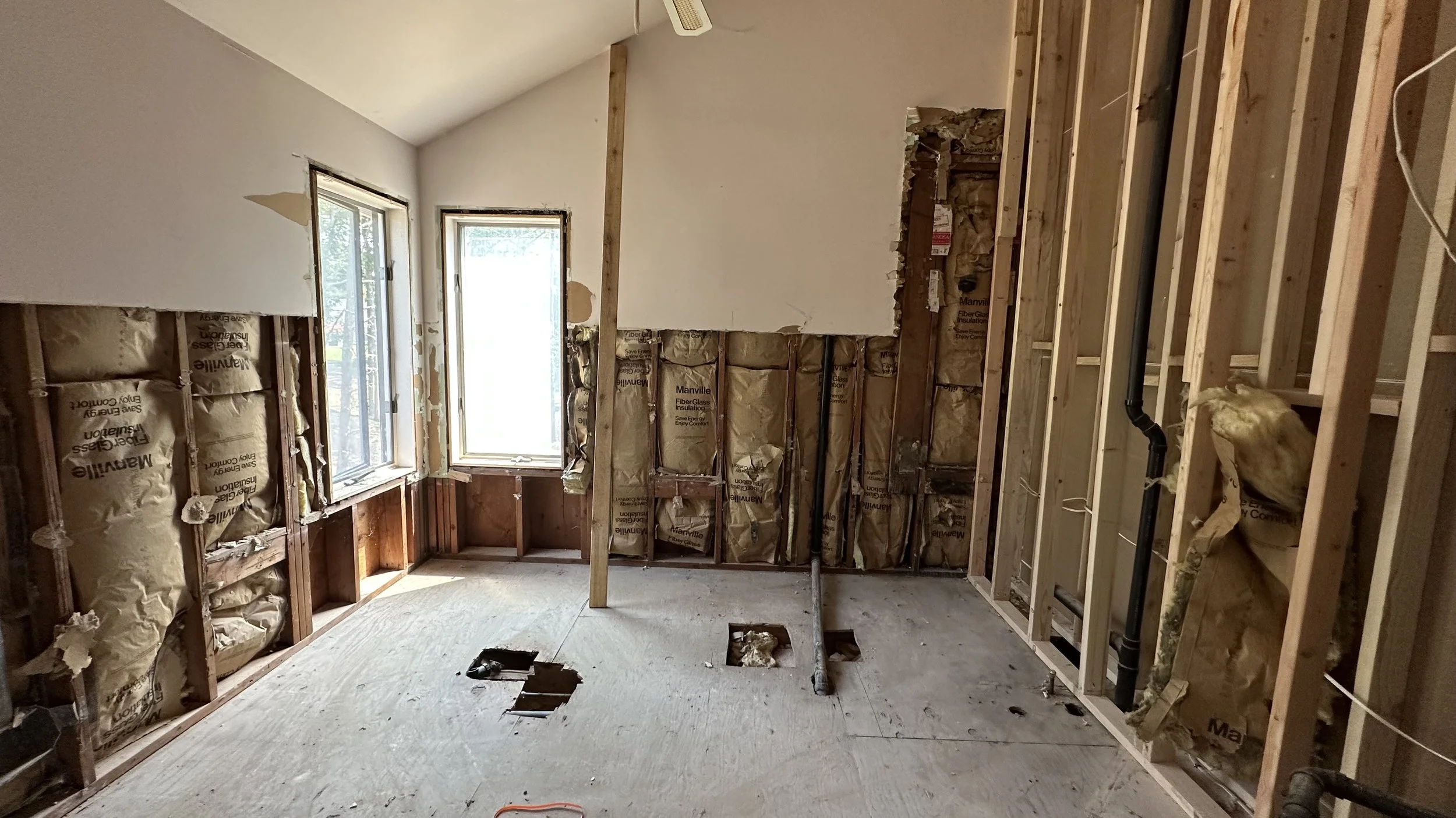 Interior of a room under renovation, with insulation in the walls, exposed framing, and two windows on the back wall.