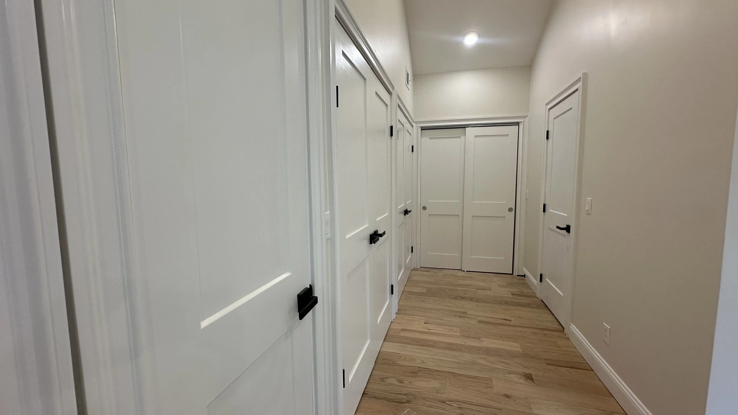 White hallway with multiple closed doors, light wood flooring, and white walls.