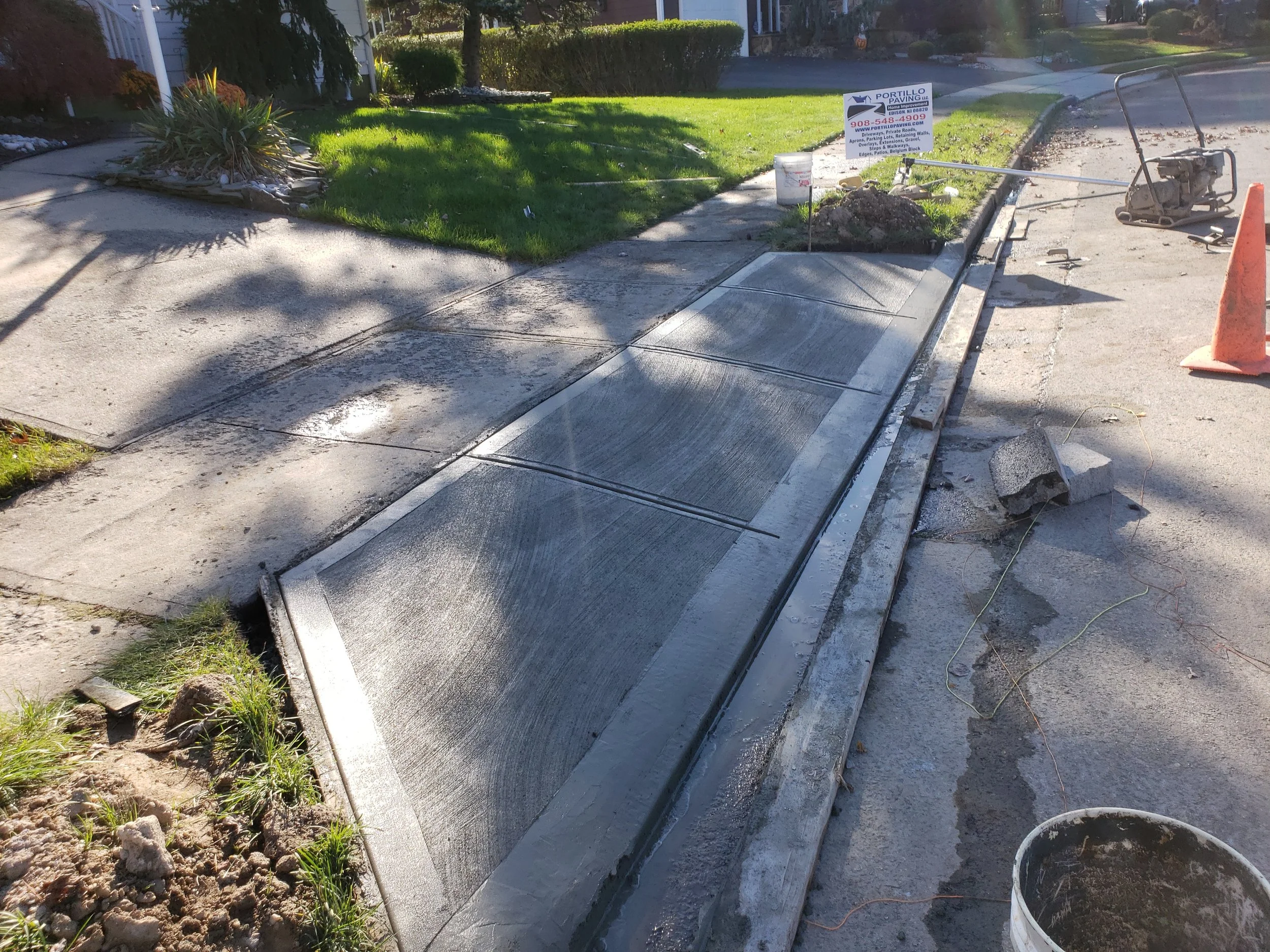 Construction workers installing a new concrete sidewalk with ramps in a residential neighborhood, with construction cones and equipment nearby.