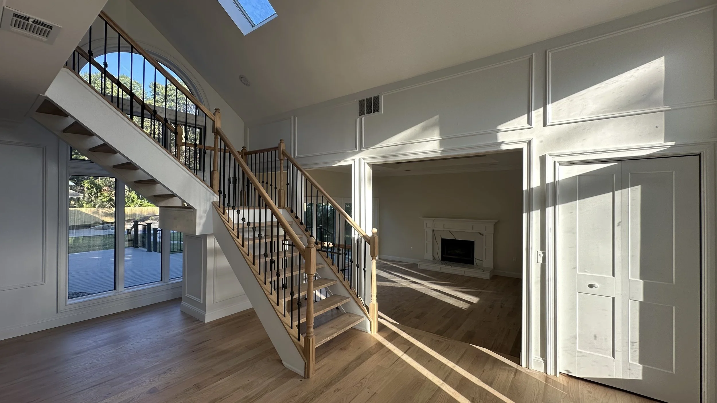 Interior view of a living room with a staircase, large windows, and sunlight casting shadows on the wall.