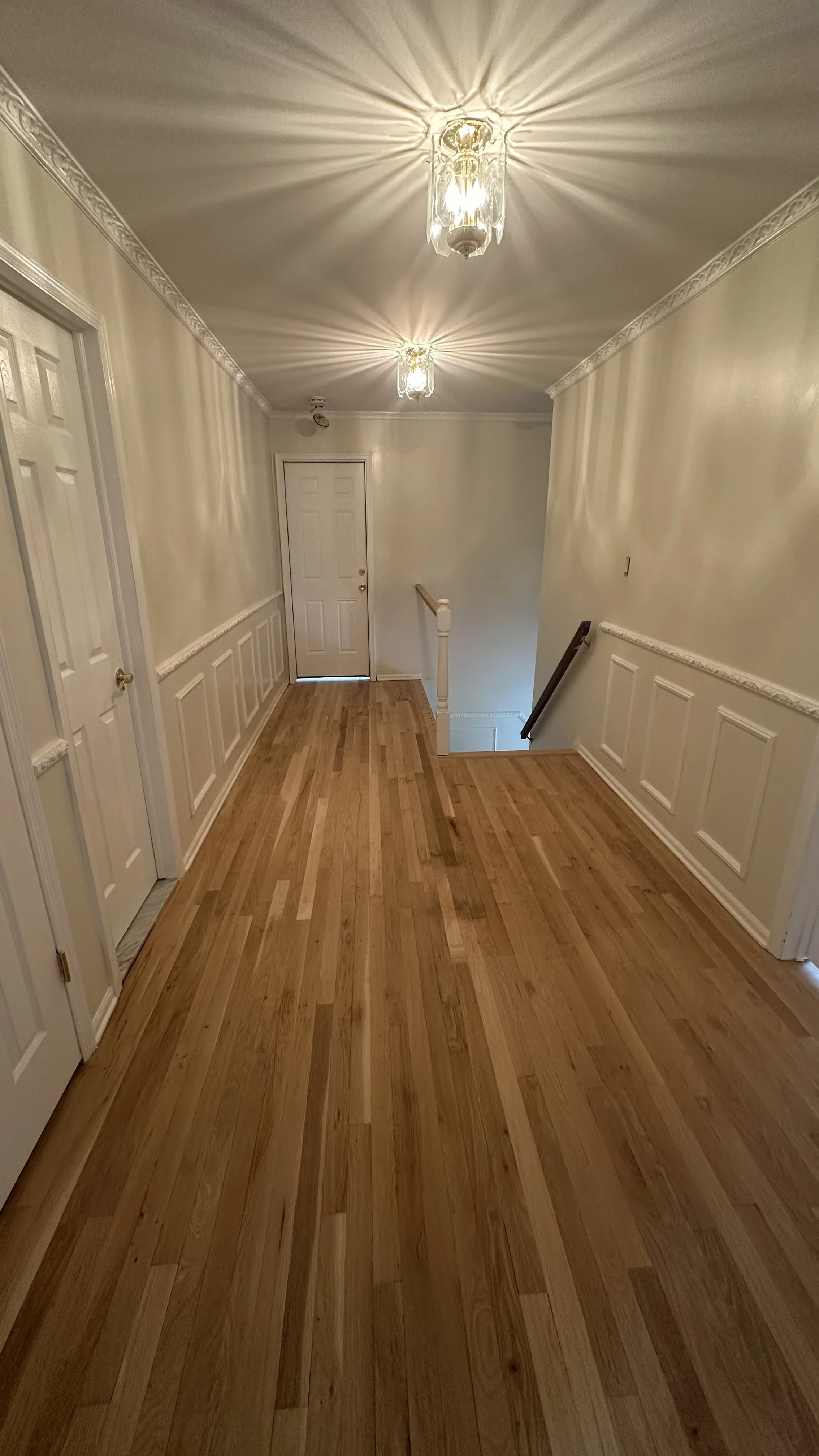 Interior of a hallway with wood flooring, white paneled walls, and two ceiling lights casting a starburst pattern, leading to a staircase and a door at the end.