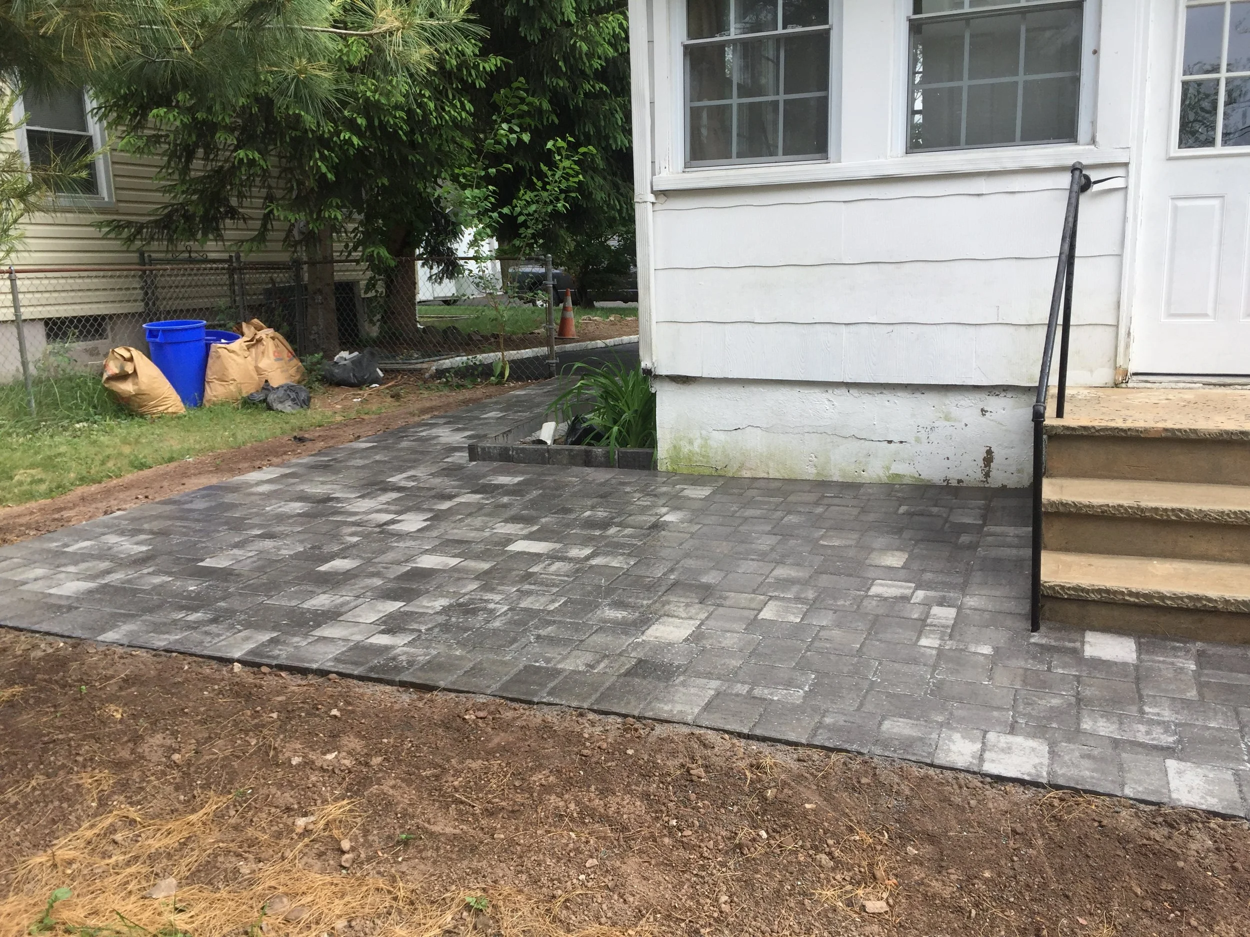 Undeveloped concrete patio area in front of a house, with stairs leading up to the entrance, adjacent to a white exterior wall with some moss and dirt.