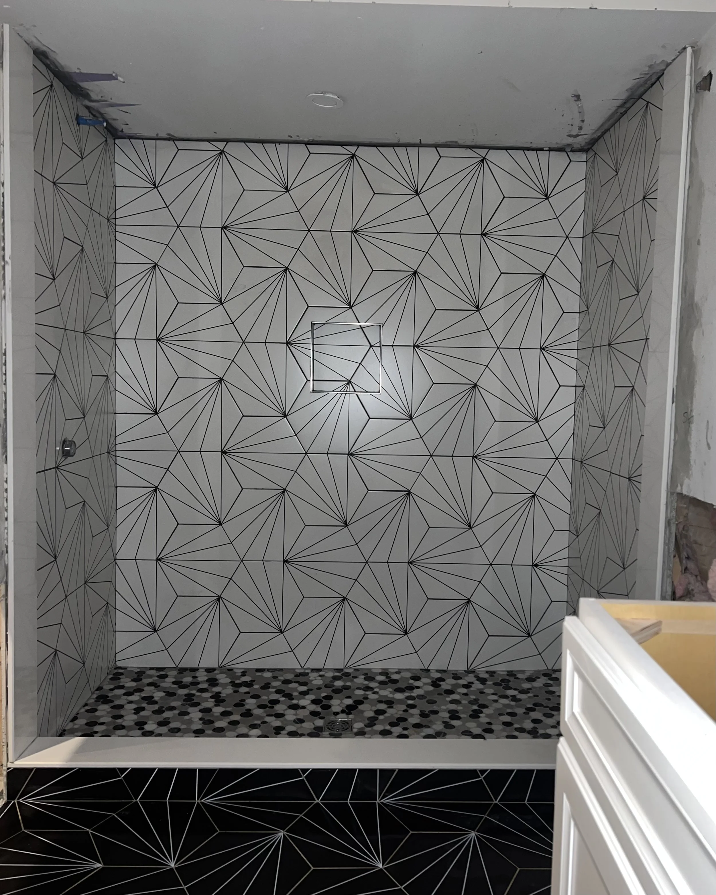 This is a bathroom shower with geometric black and white tile patterns on the walls and floor. The shower has a small square drain on the floor and a circular ceiling light fixture.