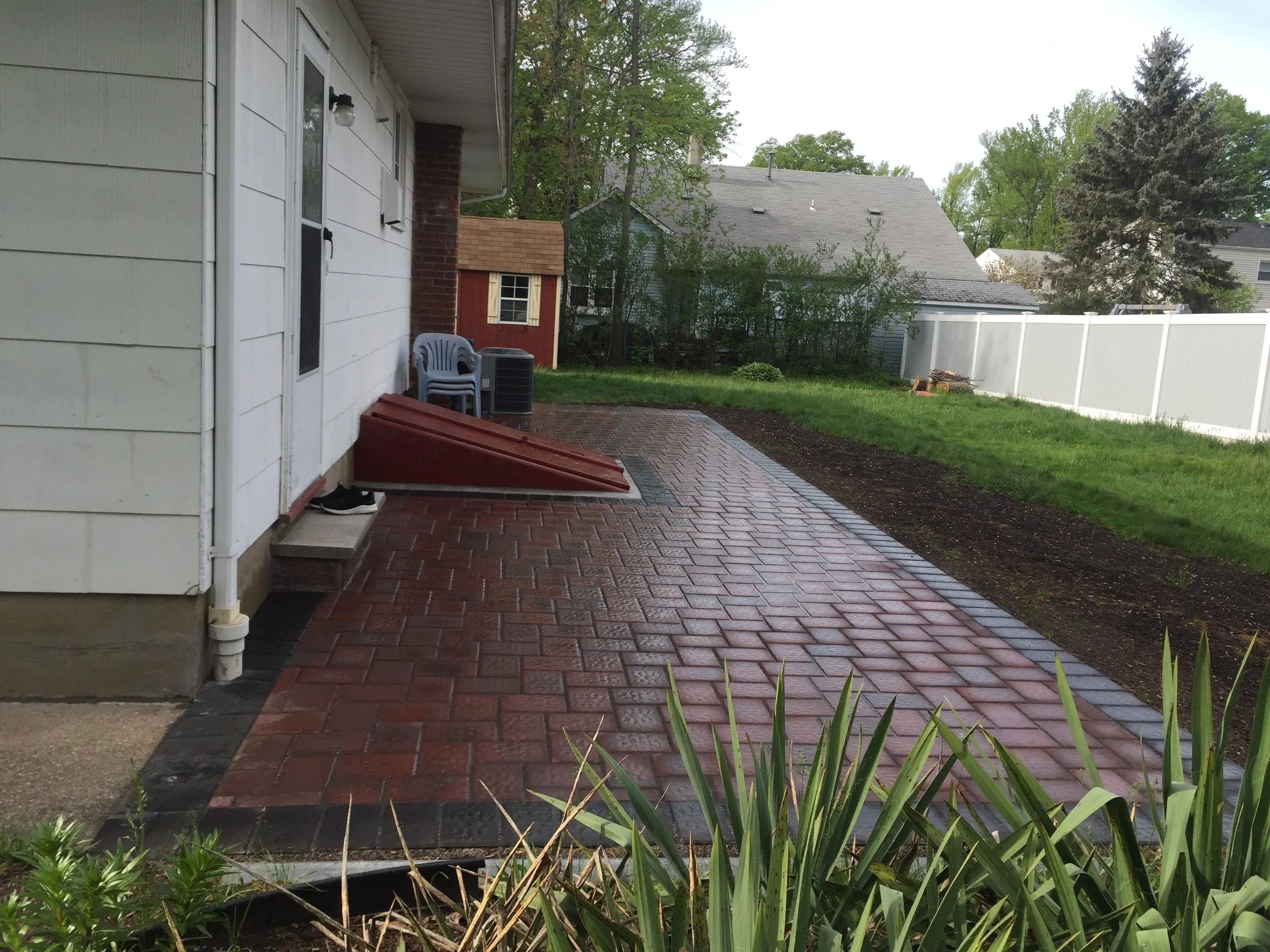 Backyard with wet red brick patio, white house wall, green grass, small shed, and trees in background.