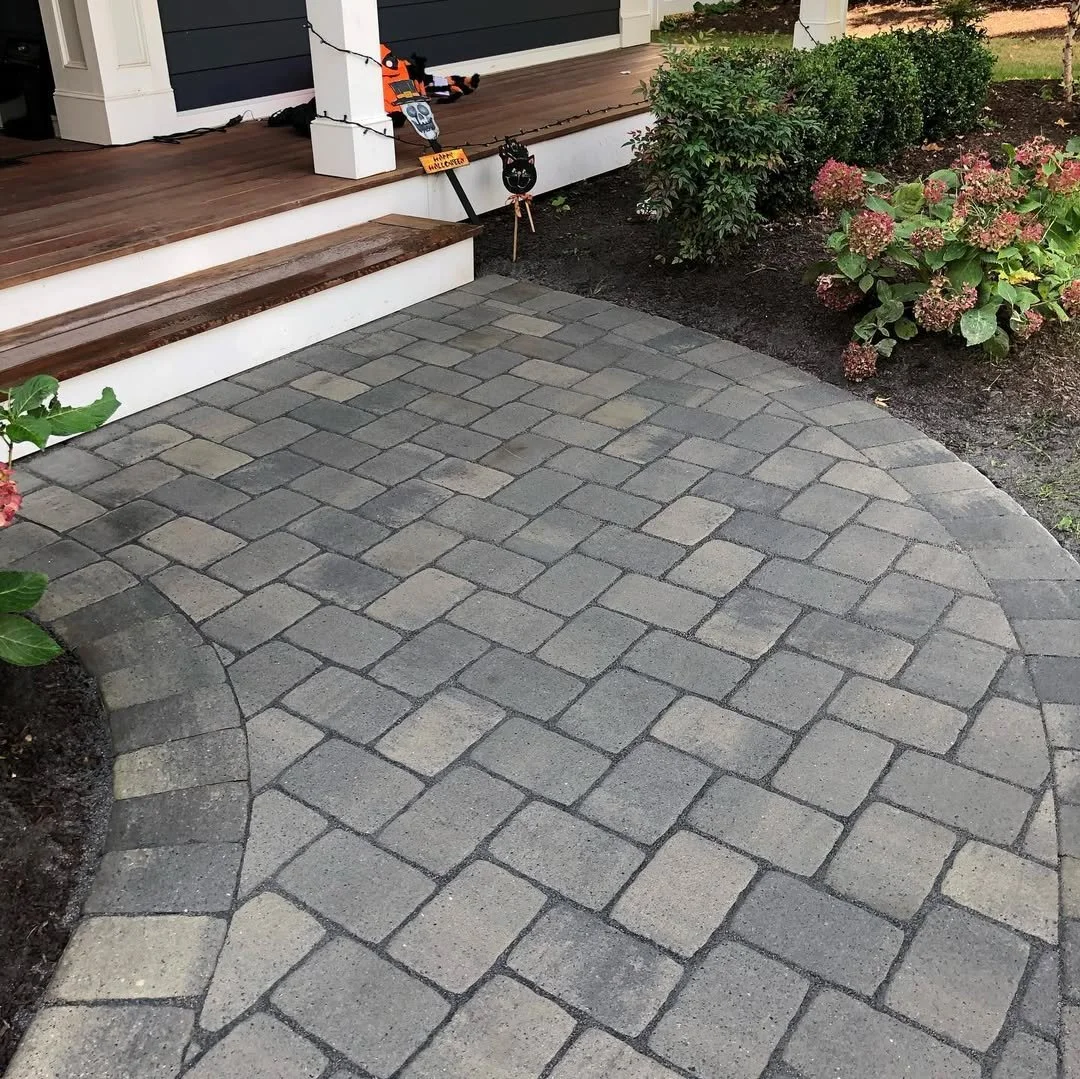Gray interlocking paver walkway leading to a house with a wooden porch decorated with fall-themed Halloween decorations. The walkway curves around flower beds with green and pink plants.