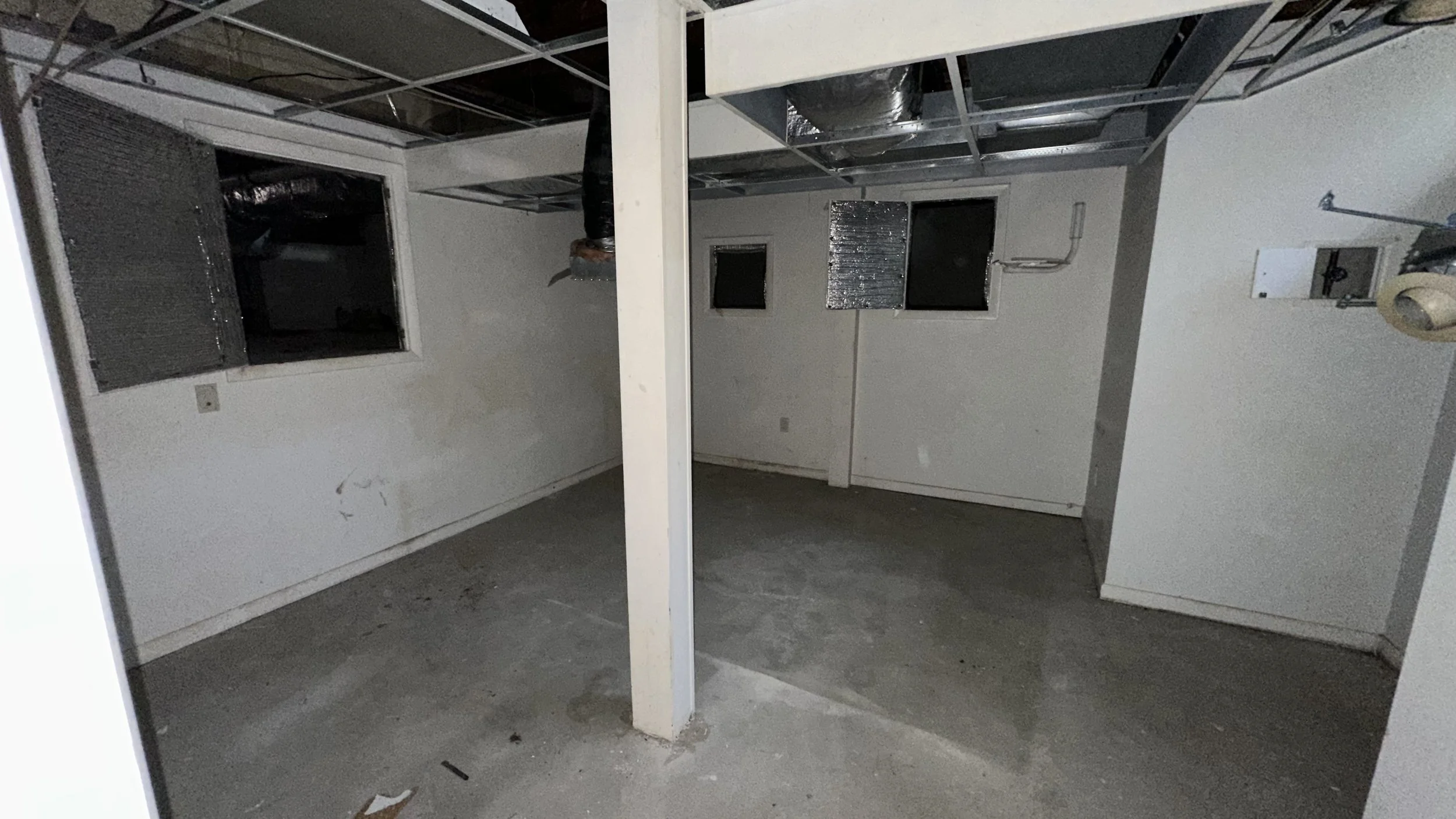 Empty basement or storage room with concrete floor, white walls, two small windows, and exposed ceiling framing.