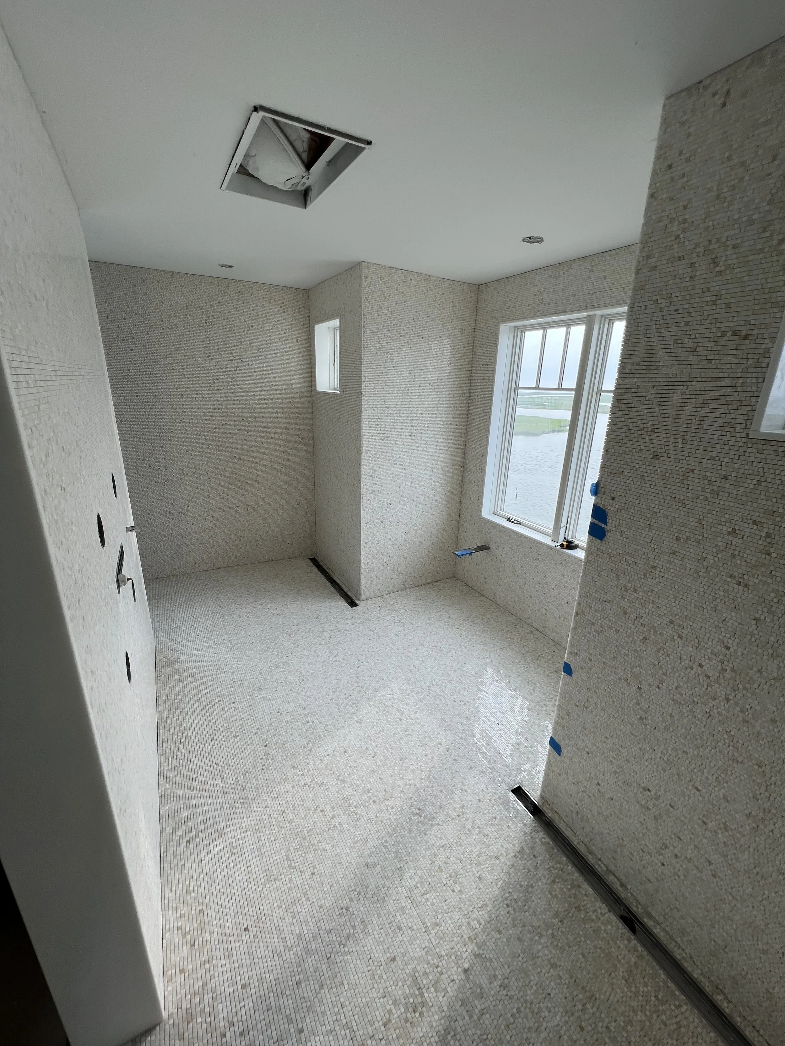 Interior of a room under construction with tiled flooring, unfinished walls, large windows, and an open ceiling hatch showing the attic space.