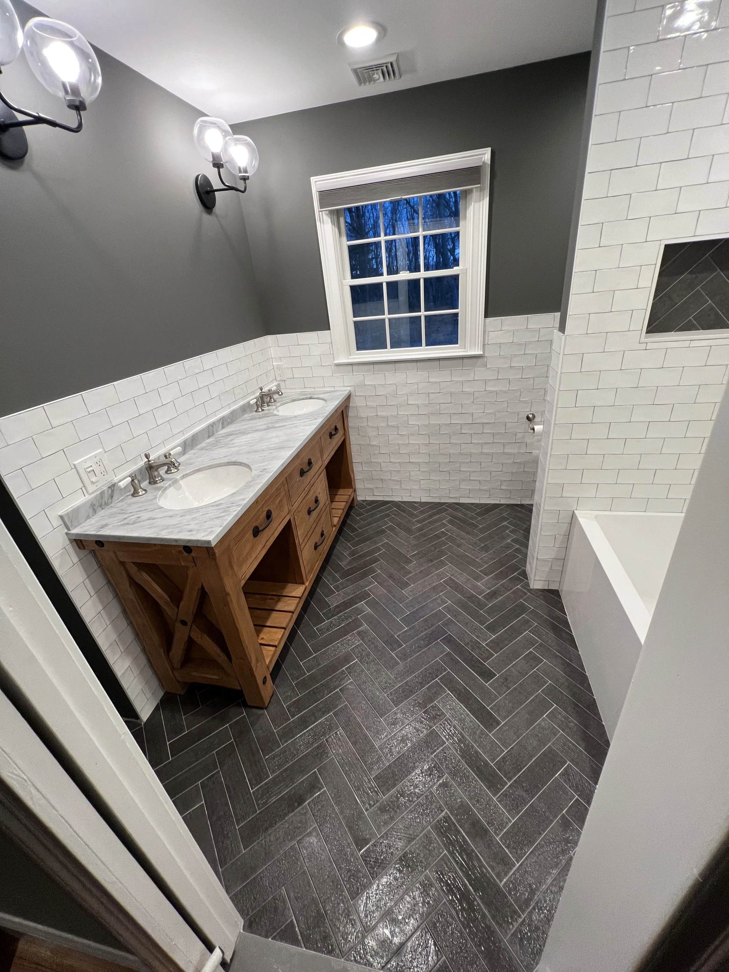 A modern bathroom with a double vanity, dark gray herringbone tile floor, white subway tile on the walls, a window with a view of trees outside, and black metal light fixtures with glass covers.