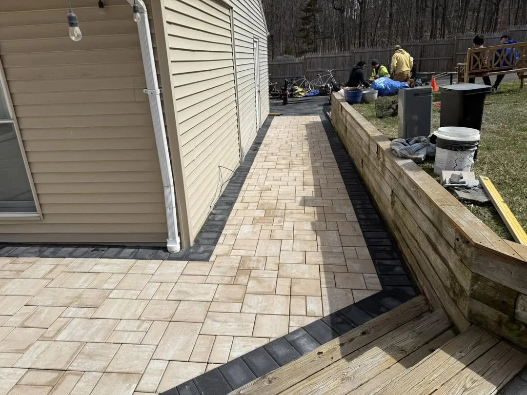 Newly paved patio with beige brick pavers and a black border beside a beige house with a white gutter. Construction materials and workers are visible in the background.