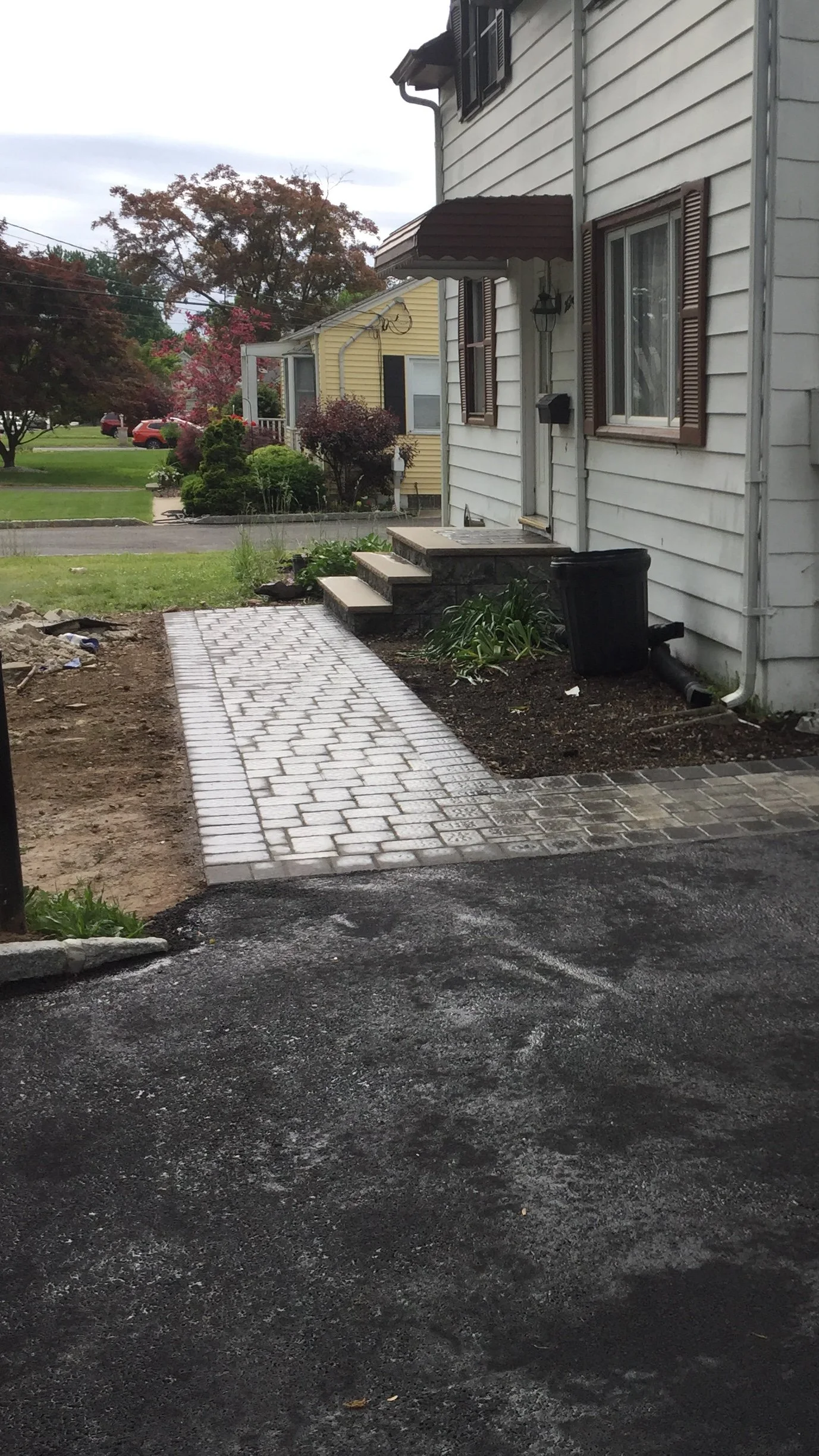 A newly paved driveway leading to a sidewalk with a brick pathway leading to house steps. The house has white siding, brown shutters, and a small porch with steps, surrounded by a landscaped yard.