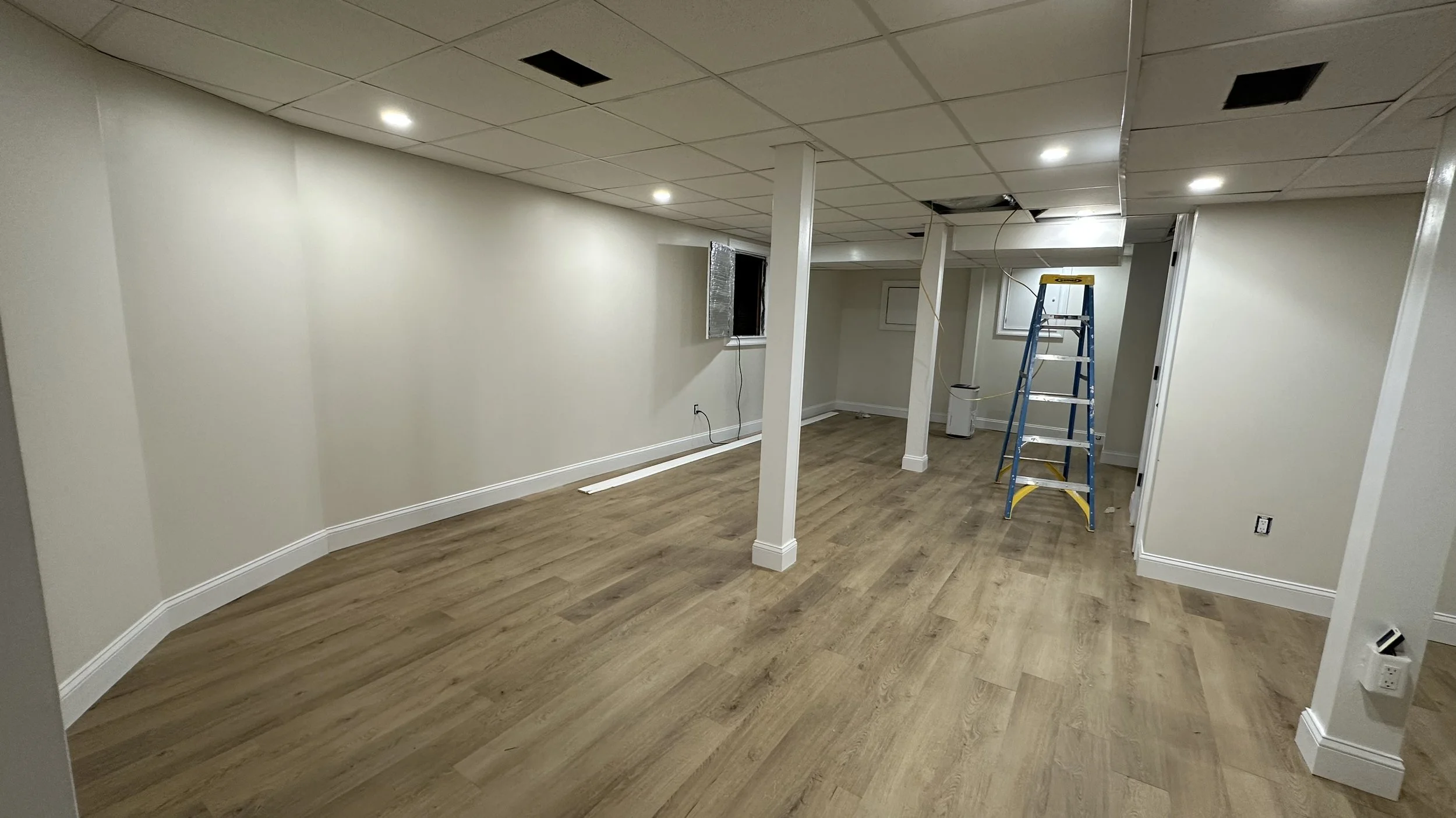 Basement room under renovation with a blue ladder, a white portable heater, and open ceiling panels showing wiring and ductwork, with light-colored hardwood floors and white walls.
