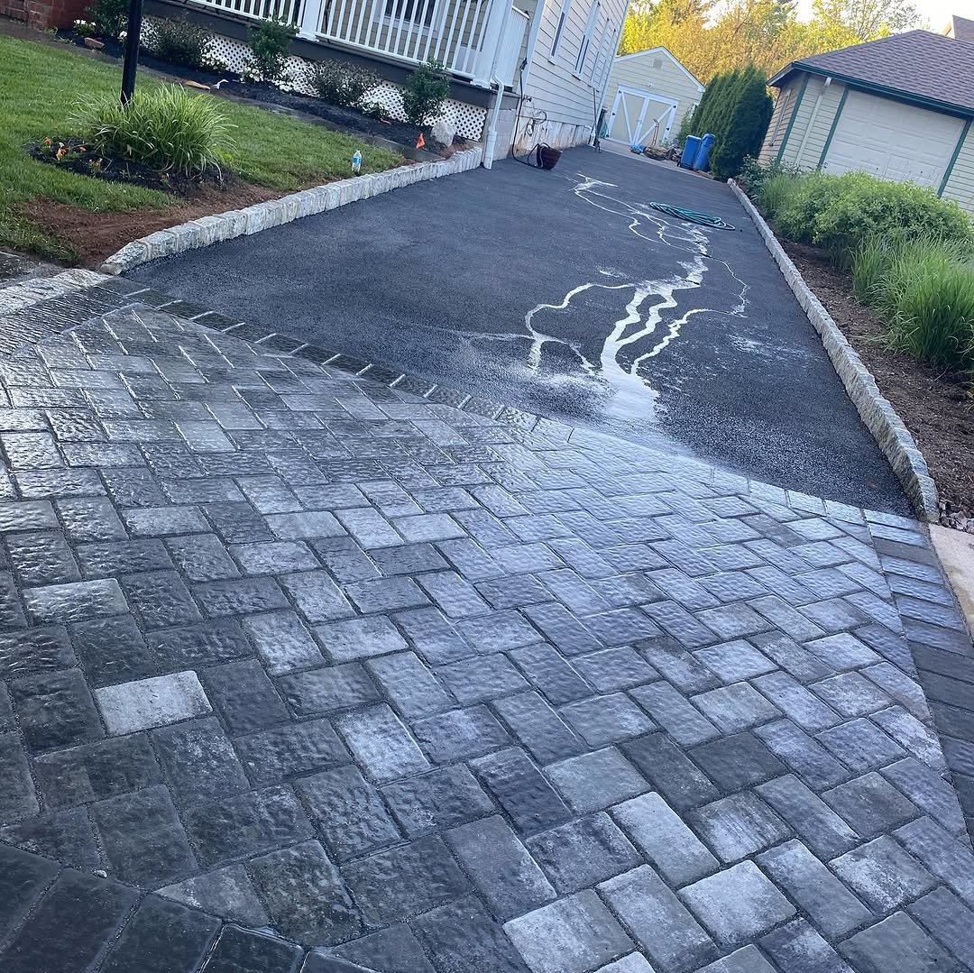 A driveway with a section of installed black paving stones and an adjacent asphalt section that has just been sealed, with excess water and sealant visible on the surface. The driveway is bordered by a brick edge and there are residential houses and 