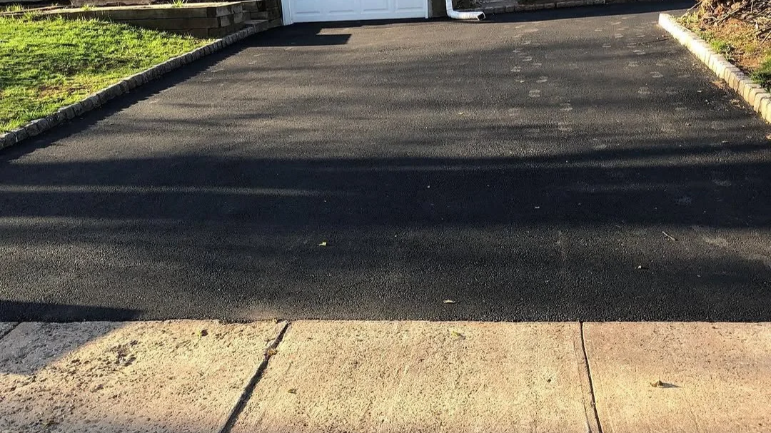 Freshly paved black asphalt driveway with a concrete sidewalk in the foreground and a grassy yard on the left, with sunlight casting shadows.