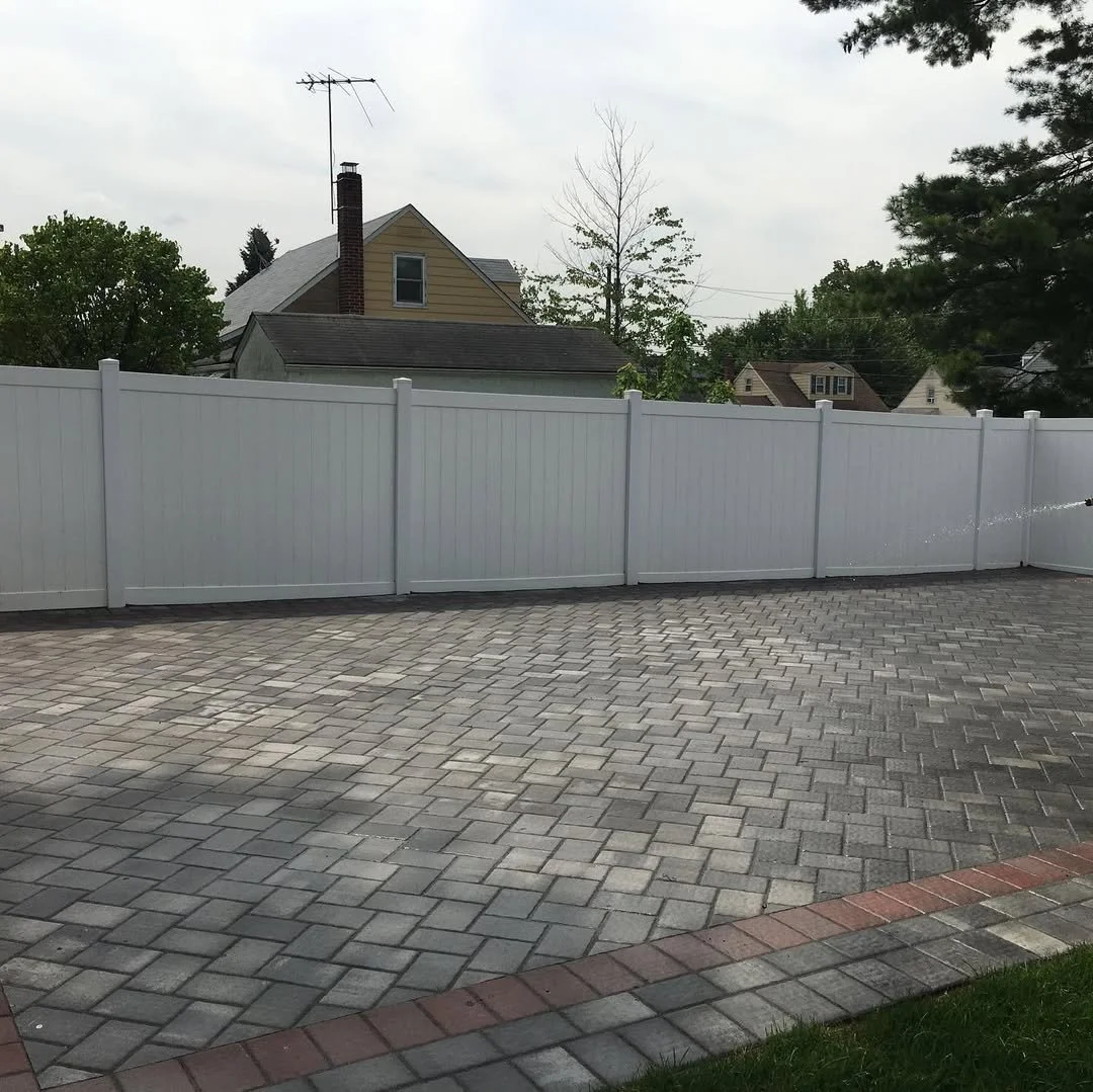 A backyard with a paved stone patio area, a white privacy fence, and houses with trees in the background.