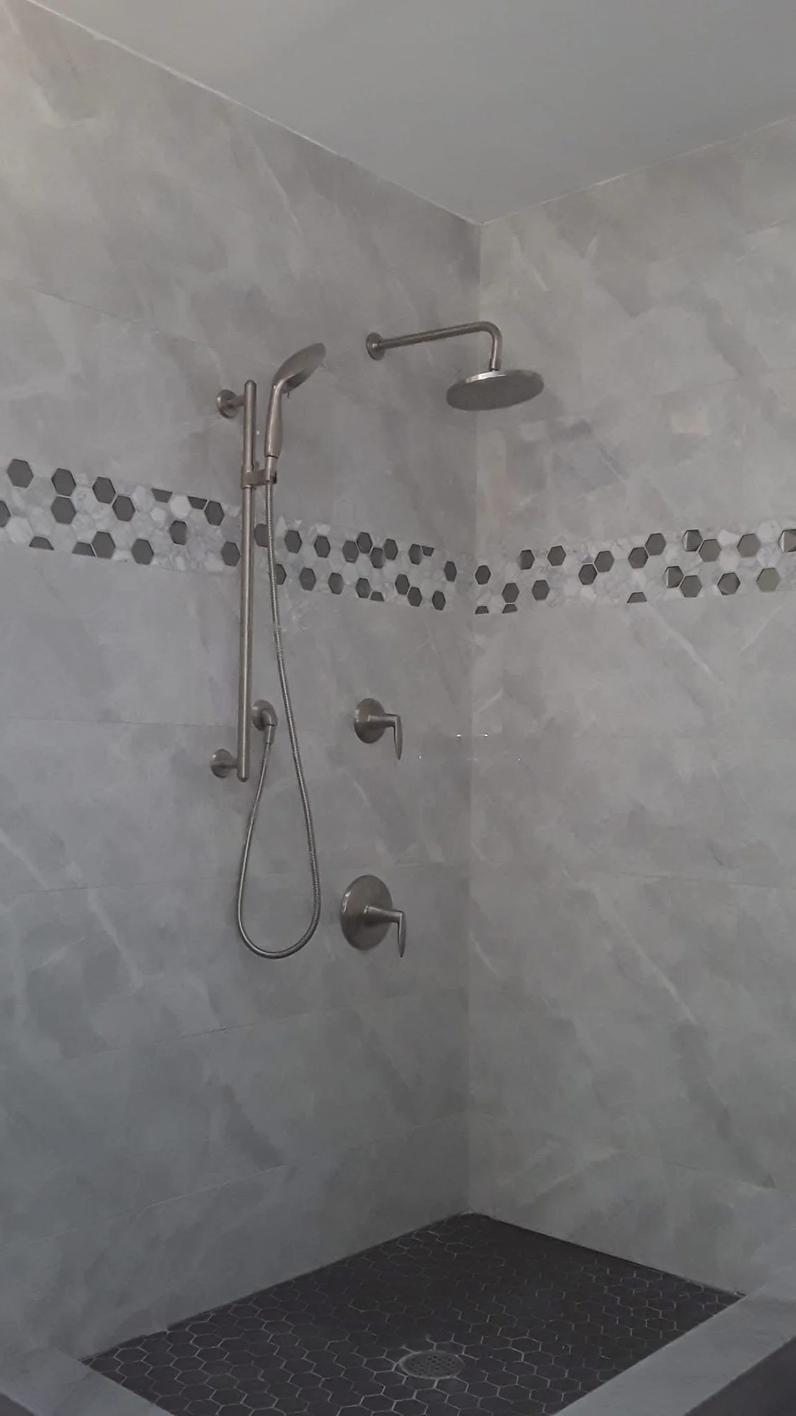 Shower area with two showerheads, one fixed and one handheld, and a grab bar, with a dark tiled floor and gray tiled walls featuring a hexagon pattern strip.