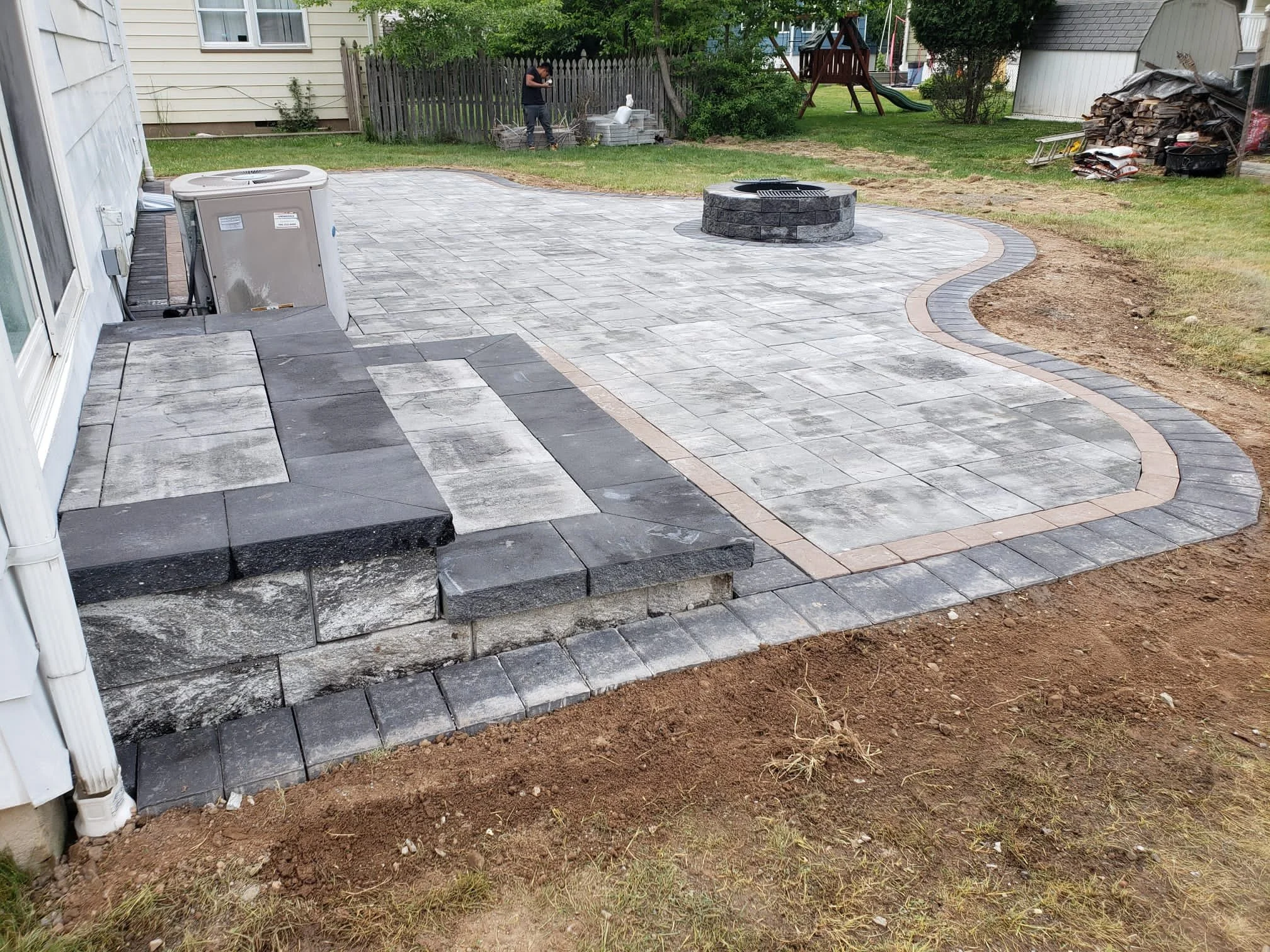 Backyard patio area with gray pavers, a curved border of bricks, a fire pit, and a small slide in the grassy yard. A person is working near some stones, and there is a pile of wood and tools in the background.
