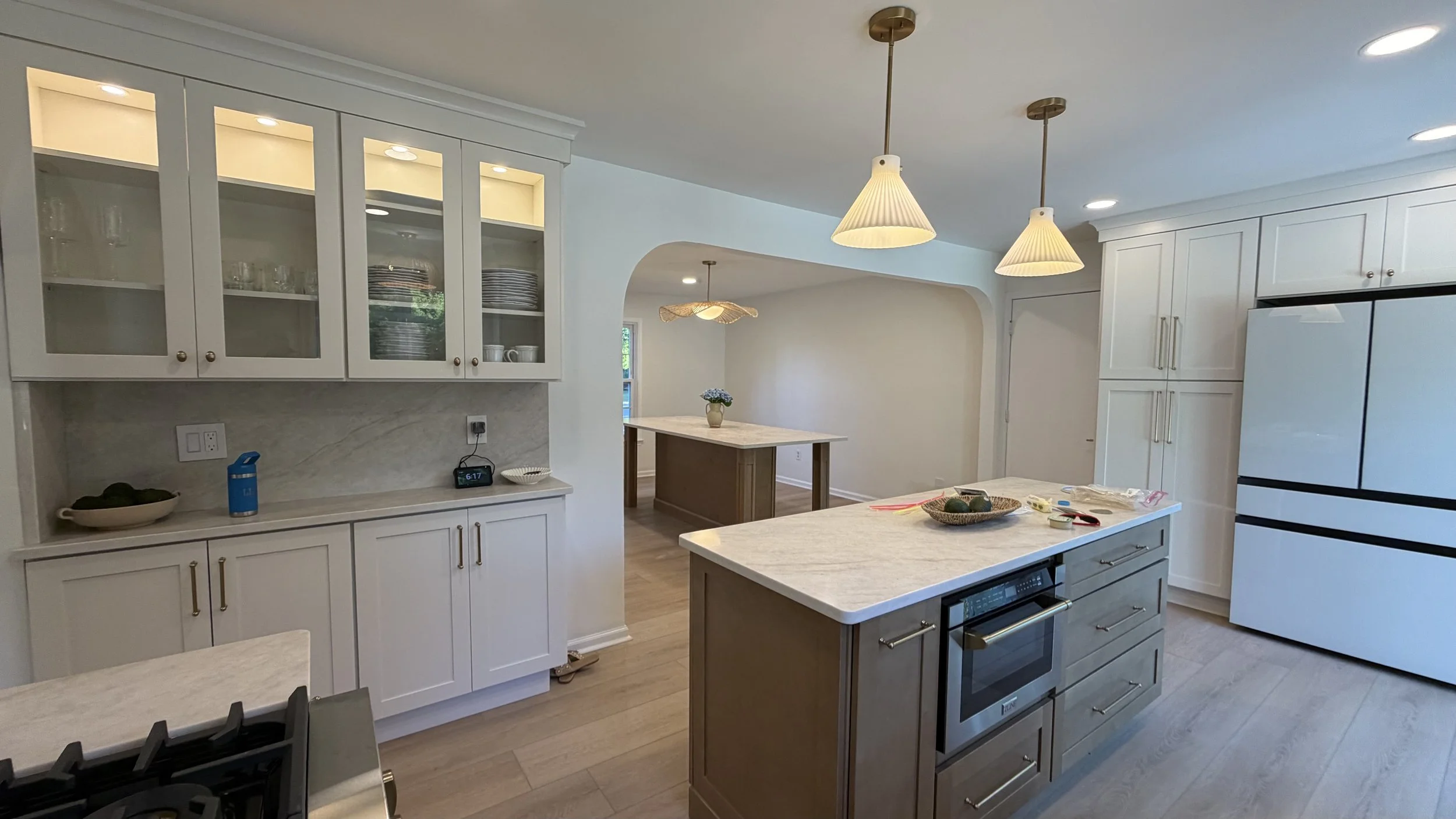 Modern kitchen with white cabinetry, marble countertops, and pendant lights. Items on the counters include a bowl of green avocados, a blue water bottle, and a digital clock. There are glass-front cabinets with dishes, and an open floor plan with a dining table and decorative lighting.