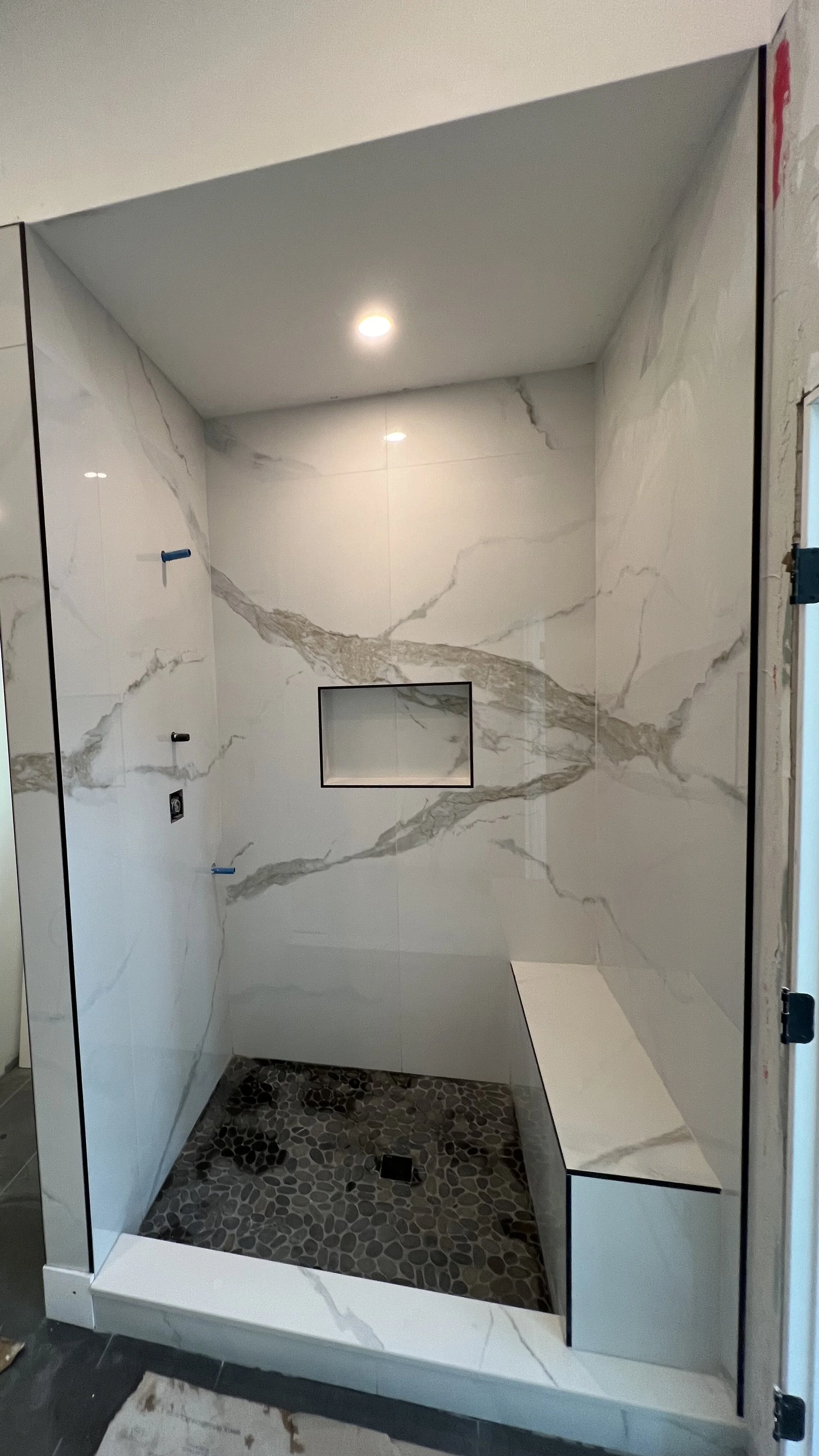 A modern walk-in shower with marble walls featuring prominent gray veining, a pebble stone shower floor, a niche built into the wall, and a glass enclosure.