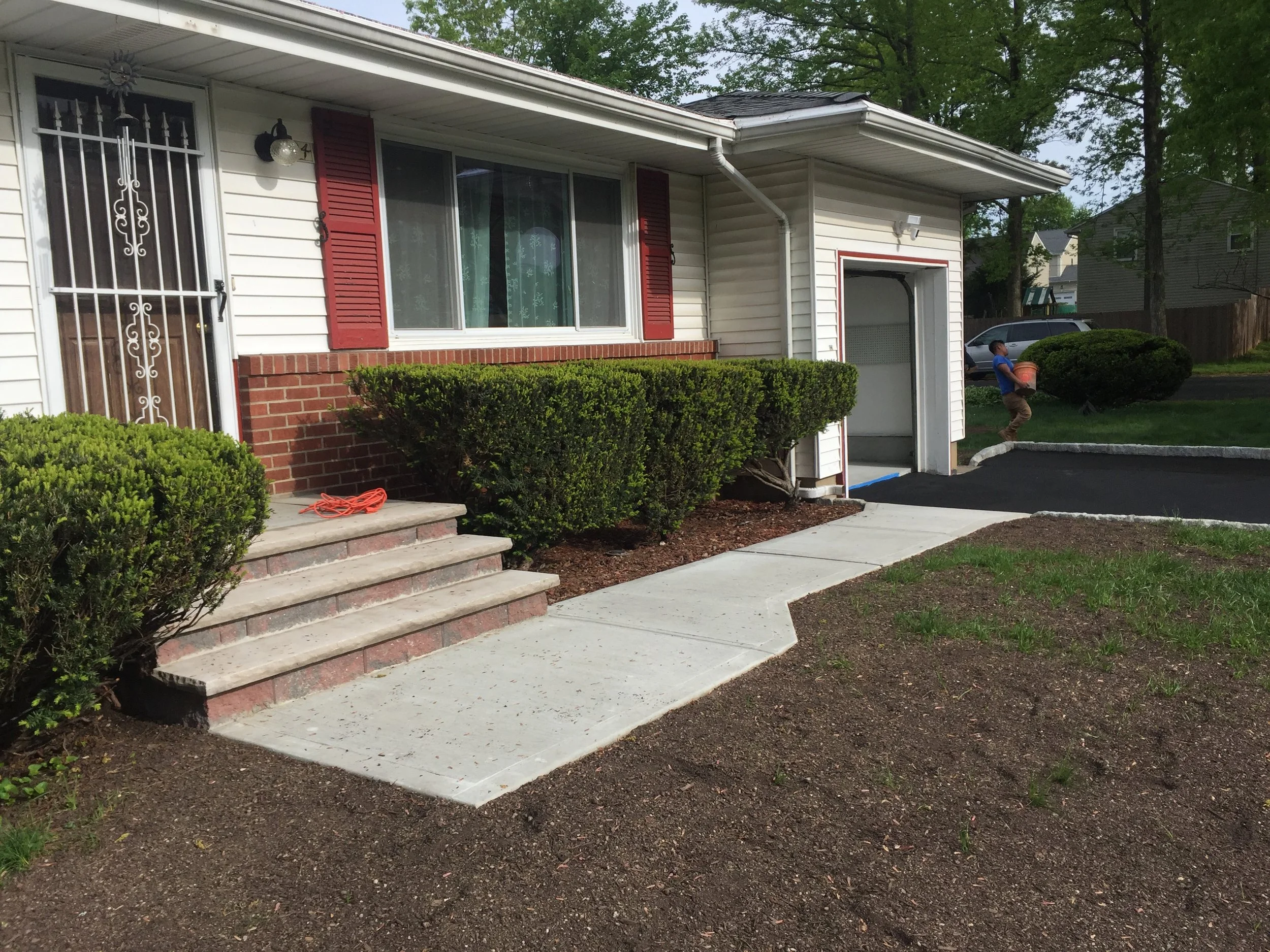 A house with white siding and red shutters, a concrete walkway leading to the front door, and a small child playing with a bucket in the yard.